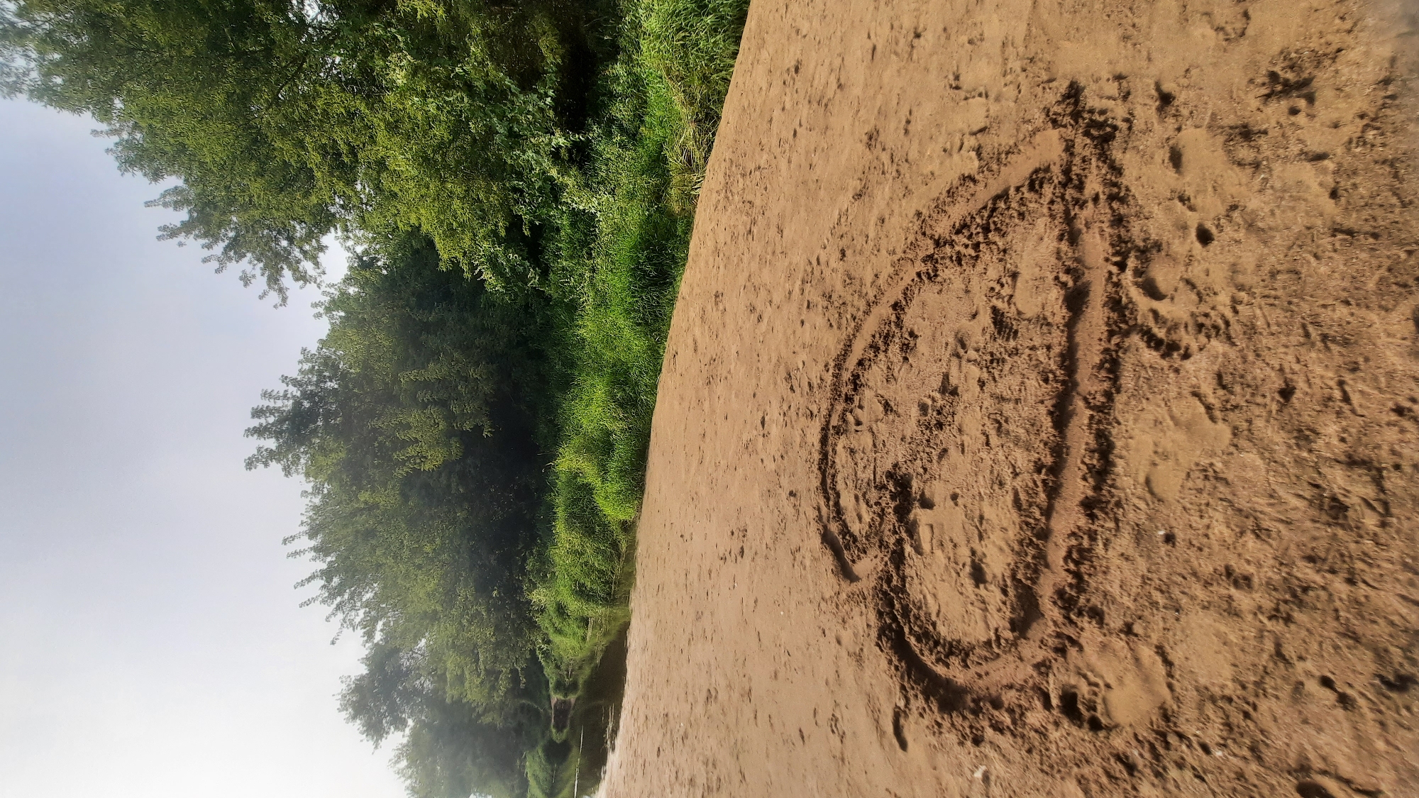 The image shows a sandy riverbank with a heart shape drawn in the sand in the foreground. The river flows to the left, bordered by lush green grass and dense trees. The sky is bright and overcast. The scene appears to be a natural landscape, likely in a rural or park setting within the vicinity of Шклоў, Belarus, given the provided context. There are no people or animals visible in the image, and no specific objects other than the heart drawing in the sand.
