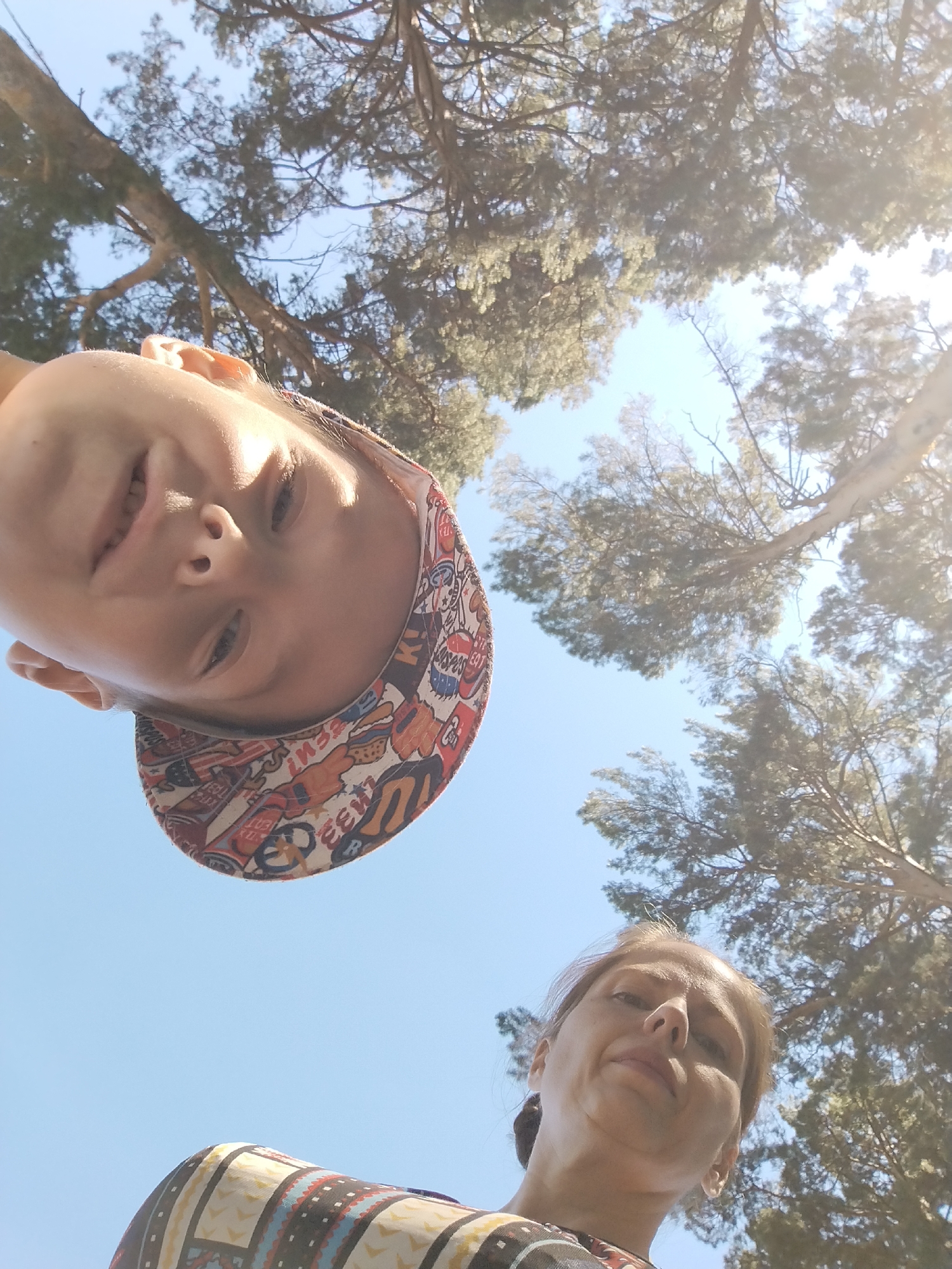A low-angle, outdoor, selfie-style photograph captures two individuals looking up towards the camera against a backdrop of trees and a clear blue sky. The person in the foreground, at the top of the frame, is a child wearing a colorful, cartoon-patterned baseball cap. The child's face is tilted downwards towards the camera, with a slight smile. Below the child, occupying the lower portion of the frame, is an adult woman. Her face is also angled upwards towards the camera, with a neutral expression. She is wearing a patterned shirt. Both individuals appear to be outdoors, possibly in a park or wooded area, with the sun creating some lens flare in the upper right corner of the image. The exact location of Zavodoukovsk, Russia is not identifiable from the visual information present in the photograph.