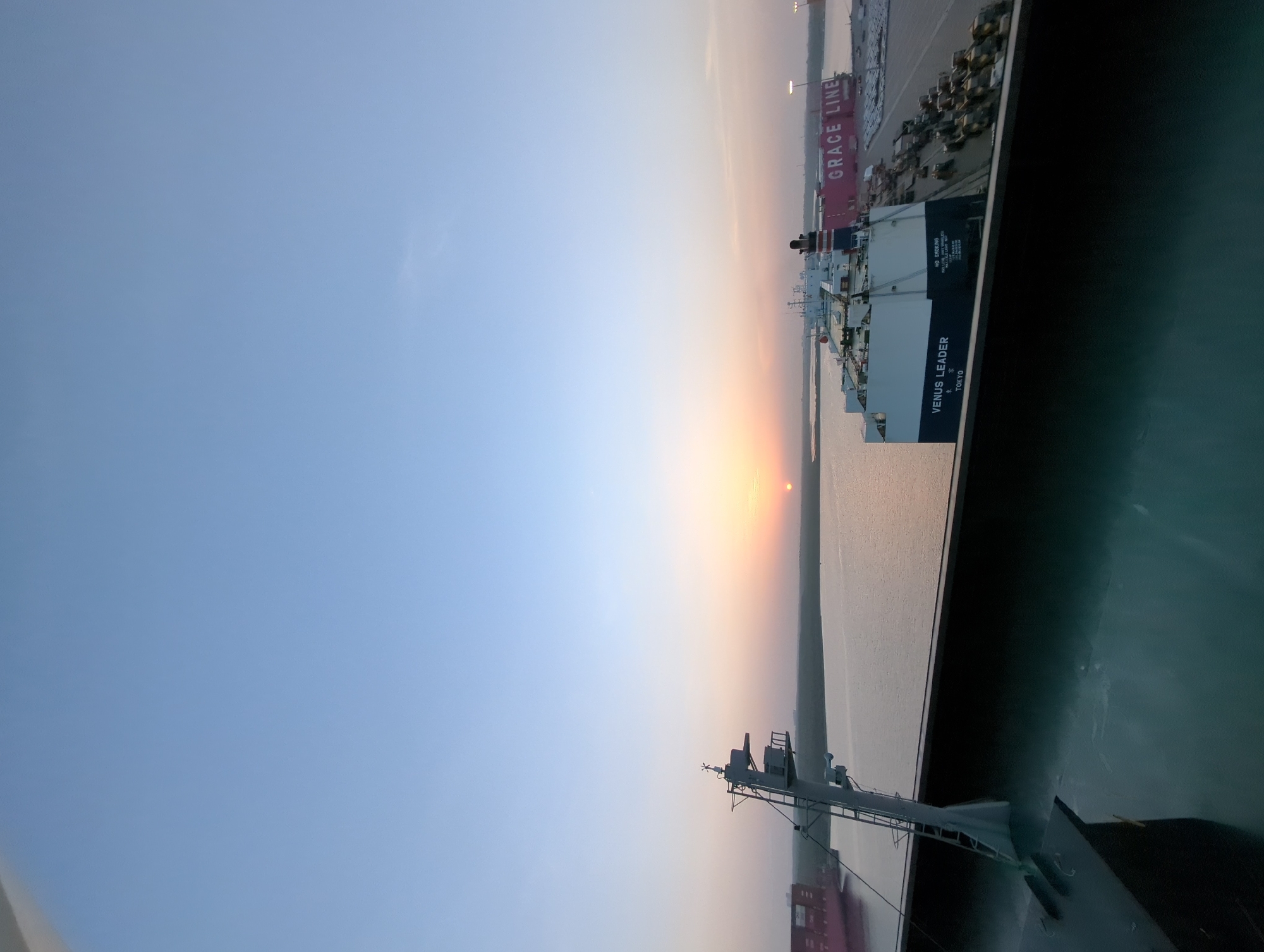The image shows a sunrise over the port of Hambantota, Sri Lanka. The sun is low on the horizon, casting a warm glow over the scene. In the foreground, the deck of a ship is visible, with a mast and some equipment. In the middle ground, the water of the port is calm, reflecting the colors of the sunrise. Several ships are docked in the port, including a large cargo ship with the name "VENUS LEADER" and "TOKYO" visible, and another vessel with "GRACE LINE" written on its side. The background shows a green, vegetated shoreline and distant structures. The sky is a clear, pale blue, with the sun creating an orange and pink gradient near the horizon. The scene is peaceful and serene, capturing the early morning light at the port.