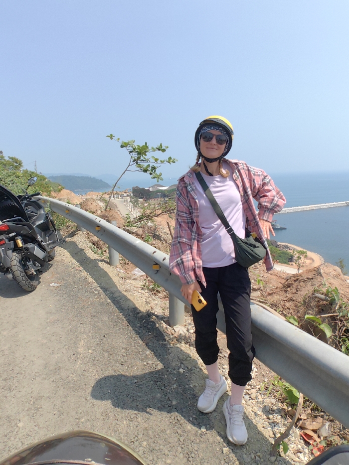 An adult female subject stands on the paved shoulder of a road, overlooking a coastal vista in Hải Vân Ward, Vietnam. The subject, positioned centrally, wears a yellow open-face helmet over a blue patterned bandana, dark sunglasses, a light purple t-shirt beneath an open pink and white plaid long-sleeve shirt, black cuffed pants, light pink socks, and white athletic shoes. A dark green cross-body bag is slung over her left shoulder. She holds a yellow smartphone vertically in her right hand, while her left hand rests on her hip. To her left, partially visible, is a parked dark grey adventure-style motorcycle. A silver metal guardrail with yellow circular reflectors spans horizontally across the mid-ground. Beyond the guardrail, a barren, rocky hillside slopes down to a developed coastal area featuring buildings, roads, and a concrete breakwater extending into the calm blue ocean. The sky above is clear and light blue. The scene is illuminated by bright sunlight, casting visible shadows.
