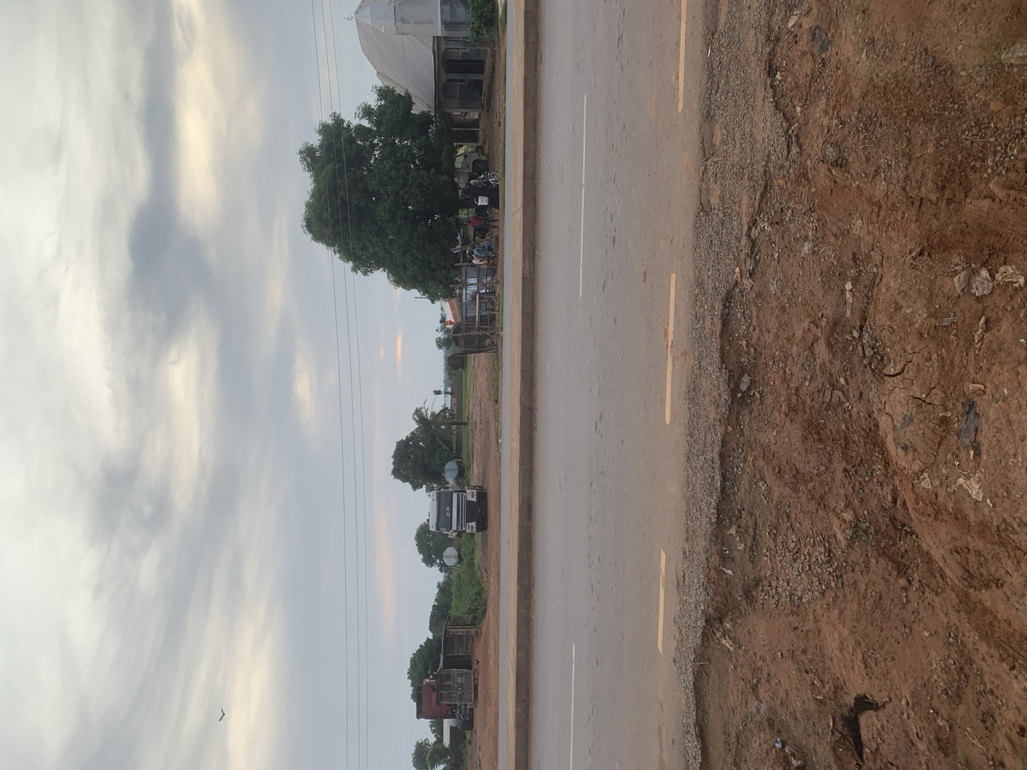 The image depicts a roadside scene in Lafia, Nigeria. In the foreground, a rough, reddish-brown dirt embankment with scattered debris lines the edge of a paved road. The road itself is marked with white and yellow lines, showing some signs of wear and tear. Across the road, several structures and vehicles are visible. To the left, there is a small, weathered wooden shelter with a few trucks and what appear to be fuel tankers parked nearby. Further to the right, a larger white truck is parked in front of a shaded area under a large tree. A group of people are gathered near this area, and a bicycle is visible. Beyond these structures, a light-colored, domed building with an adjacent open-air section stands, possibly a place of worship. The background is filled with trees and vegetation, with a cloudy sky overhead. A bird is seen in flight in the upper left quadrant of the sky. The overall impression is of a somewhat unkempt but active roadside location in a semi-rural or peri-urban area.