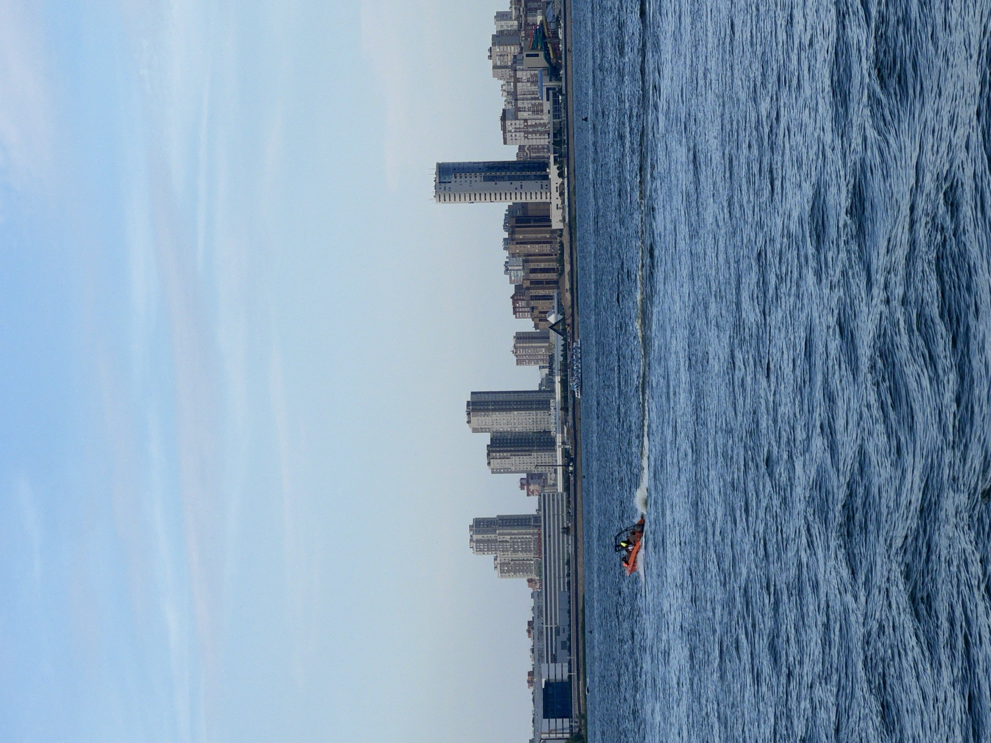 An aerial view captures a serene urban waterfront with a clear sky. The foreground is dominated by the shimmering blue waters of a river, with gentle ripples creating a dynamic texture. In the mid-ground, an orange jet ski, with two occupants visible, speeds across the water, leaving a white wake in its path. Beyond the river, the Kazan skyline unfolds, a panorama of modern architecture. Tall, slender skyscrapers, interspersed with lower-rise buildings and a large shopping center, form a striking silhouette against the horizon. The city appears to be a vibrant hub, with a blend of residential and commercial structures. The overall scene depicts a harmonious coexistence of nature and urban development.