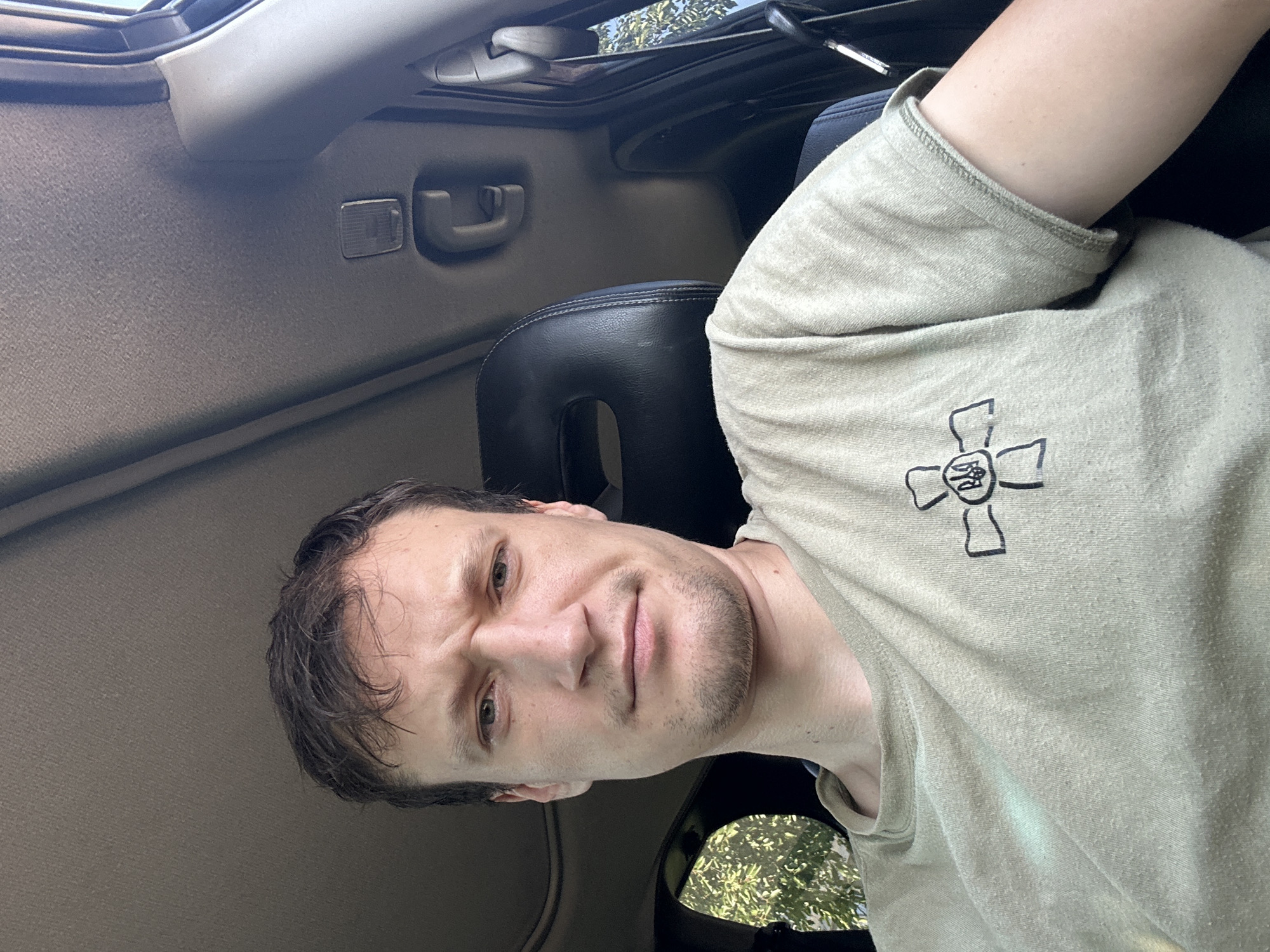 A man with dark hair and a short beard, wearing a light tan t-shirt with a black emblem of a cross with a trident in the center, sits in the driver's seat of a vehicle. He is looking forward with a slightly furrowed brow. The interior of the vehicle, including the headrest of the passenger seat and parts of the car's ceiling, are visible. Sunlight streams in from outside, illuminating the man and the car's interior. The man's t-shirt has a textured appearance due to pilling. Trees and foliage are visible through the window on the left side of the frame. The image appears to have been taken during the daytime.