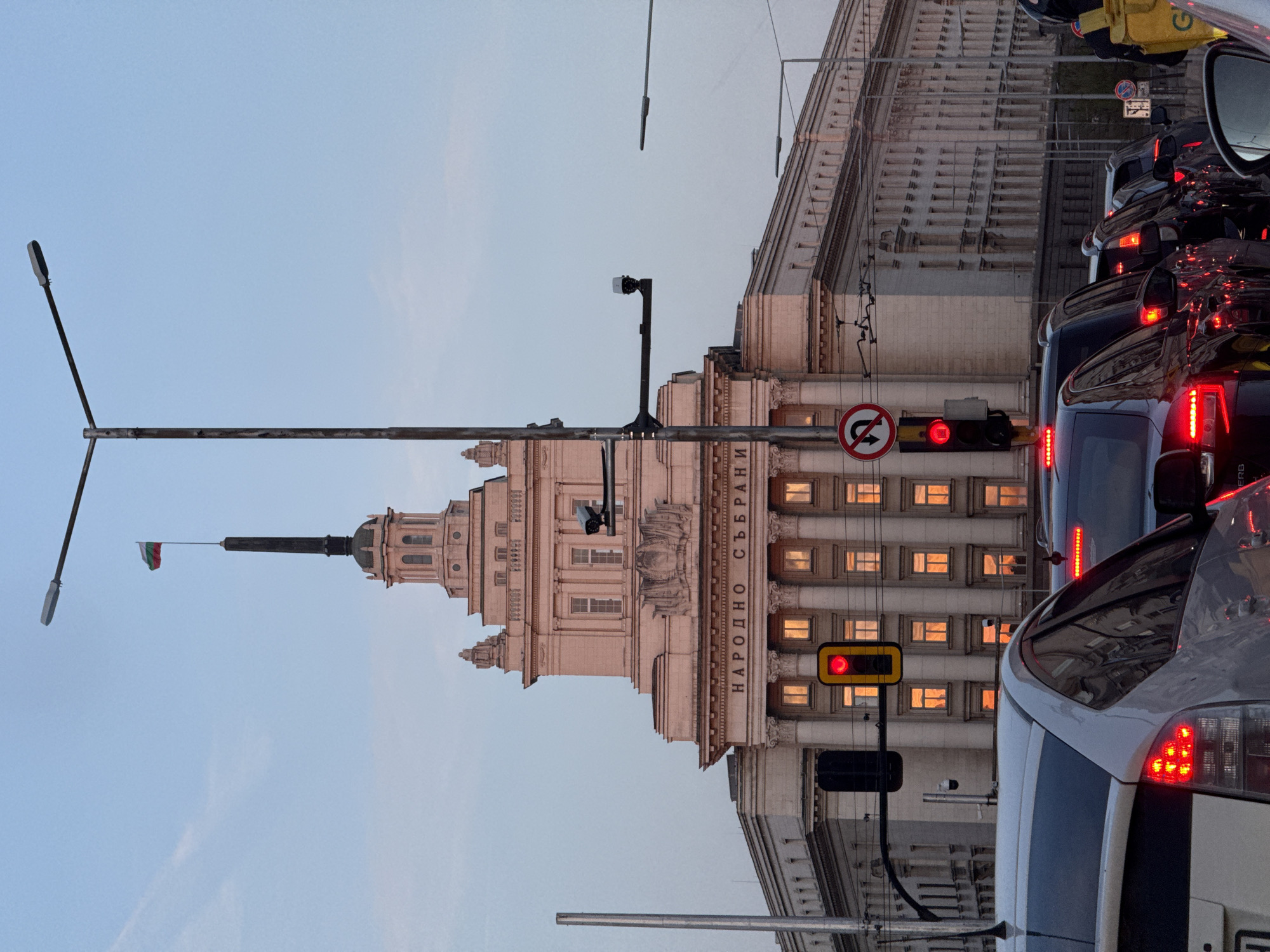 The image captures an urban street scene in Sofia, Bulgaria, during what appears to be late afternoon or early evening. The prominent subject is a grand, ornate building with a neoclassical or Stalinist-era architectural style, featuring a tall central tower topped with a spire. A Bulgarian flag, distinguishable by its green, white, and red horizontal stripes, flies from the spire. Inscribed on the building's facade, the Cyrillic text "НАРОДНО СЪБРАНИЕ" is clearly visible, identifying it as the National Assembly of Bulgaria. Many windows on the building's lower floors are illuminated with a warm, amber light, contrasting with the fading daylight.
In the foreground and midground, a line of cars is visible in traffic, occupying the right side of the frame. Their red brake lights are illuminated, suggesting they are either stopped or moving slowly. A red traffic light is prominently displayed, further indicating stopped traffic. A circular traffic sign prohibiting U-turns is also present. Several street lights and utility lines crisscross the upper parts of the scene against a clear, light blue sky. The setting is a bustling city environment, characterized by both impressive architecture and routine urban congestion.
