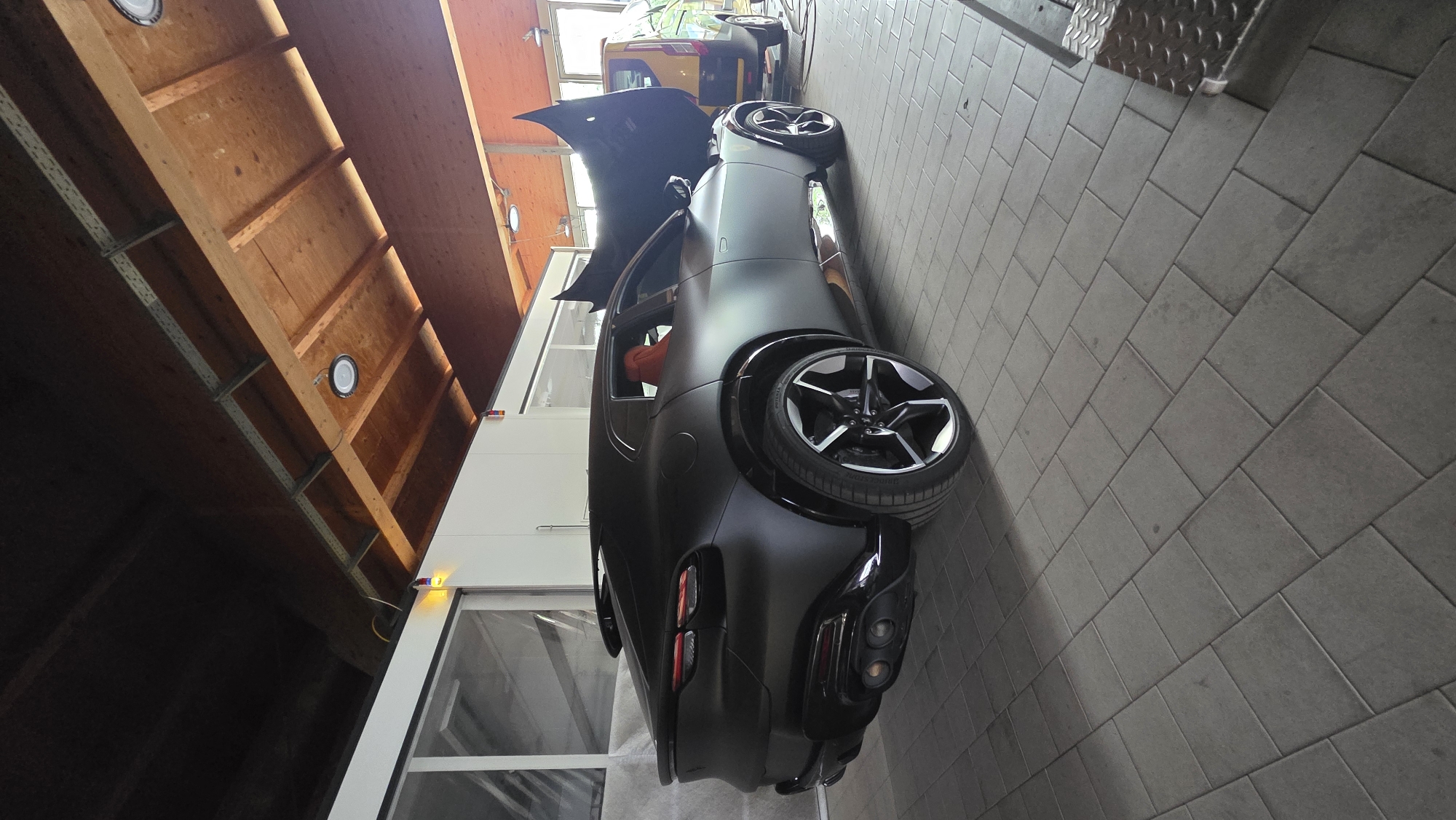 The image shows the rear and side of a matte black, four-door coupe with a red interior, parked in an indoor bay. The car's hood is open, revealing the engine compartment. The bay has a tiled floor and a wooden ceiling with exposed beams. Outside the bay, a yellow van is visible. The setting appears to be a car service or detailing facility.