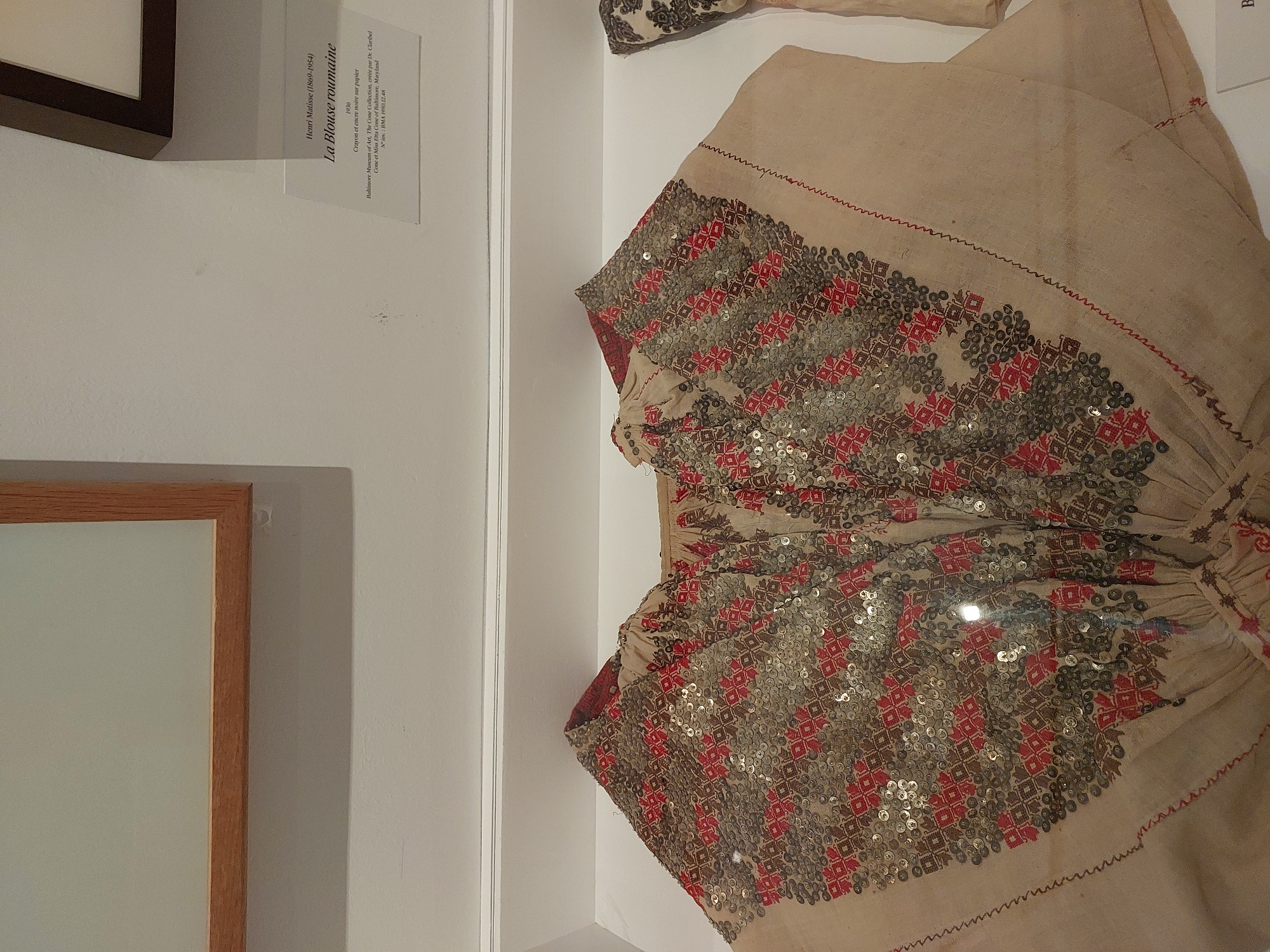 An embroidered Romanian blouse, created by Henri Matisse, is displayed behind glass in a museum. The blouse is made of beige fabric and is heavily decorated with red and brown geometric patterns and sequins. The neckline is gathered, and the sleeves are wide. A label next to the blouse reads "Henri Matisse (1869-1954) 1936 La Blouse roumaine Crayon et encre noire sur papier Baltimore Museum of Art. The Cone Collection, created par Dr. Claribel Cone et Miss Etta Cone of Baltimore, Maryland N.Y. 1984. 1984.12.24". The background is a white wall with a wooden frame visible at the top left. The setting appears to be a museum in Nice, France, given the reference to "N.Y." potentially indicating New York or a French equivalent, and the known presence of Matisse works in French museums.