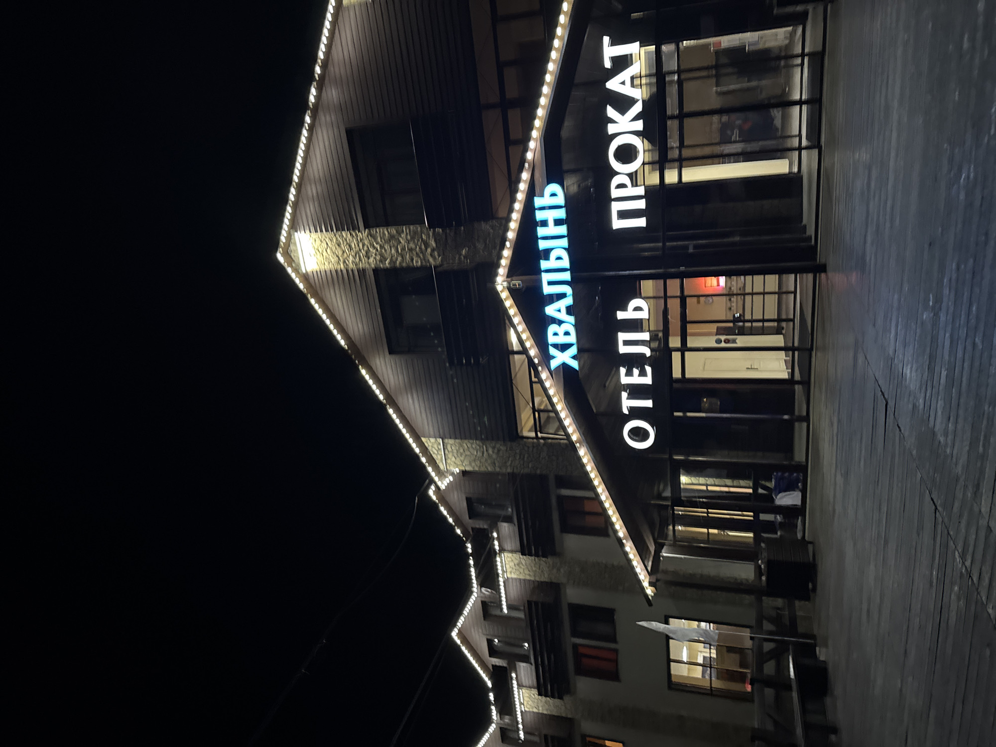 The image depicts the exterior of the "Khvalynskiy" hotel in Khvalynsk, Russia at night. The building is decorated with string lights along the roofline. A large sign attached to the building reads "ОТЕЛЬ ХВАЛЫНЬ ПРОКАТ" in vertically-oriented, illuminated letters. The building has multiple stories and a combination of textured and smooth exterior surfaces. The ground in front of the building appears to be made of wooden planks. There are benches and other items placed along the walkway.