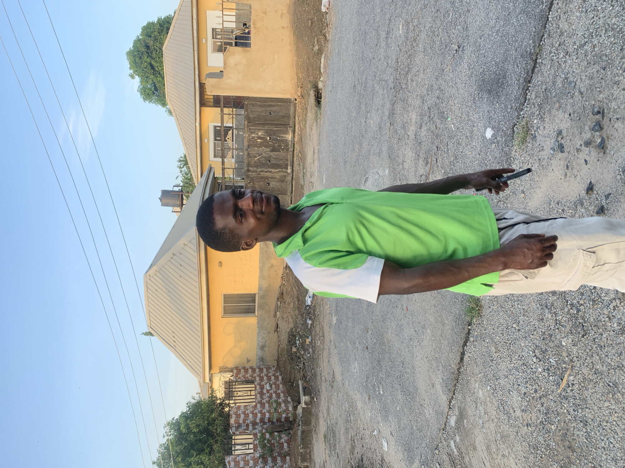A man stands on a paved area with gravel along the edges, holding a mobile phone. He is wearing a green and white polo shirt and light-colored pants. Behind him are two yellow buildings with metal roofs. The environment suggests a residential area, possibly in Abuja, Nigeria, given the description provided. Power lines are visible in the background against a blue sky. There is litter present near the base of one of the buildings.