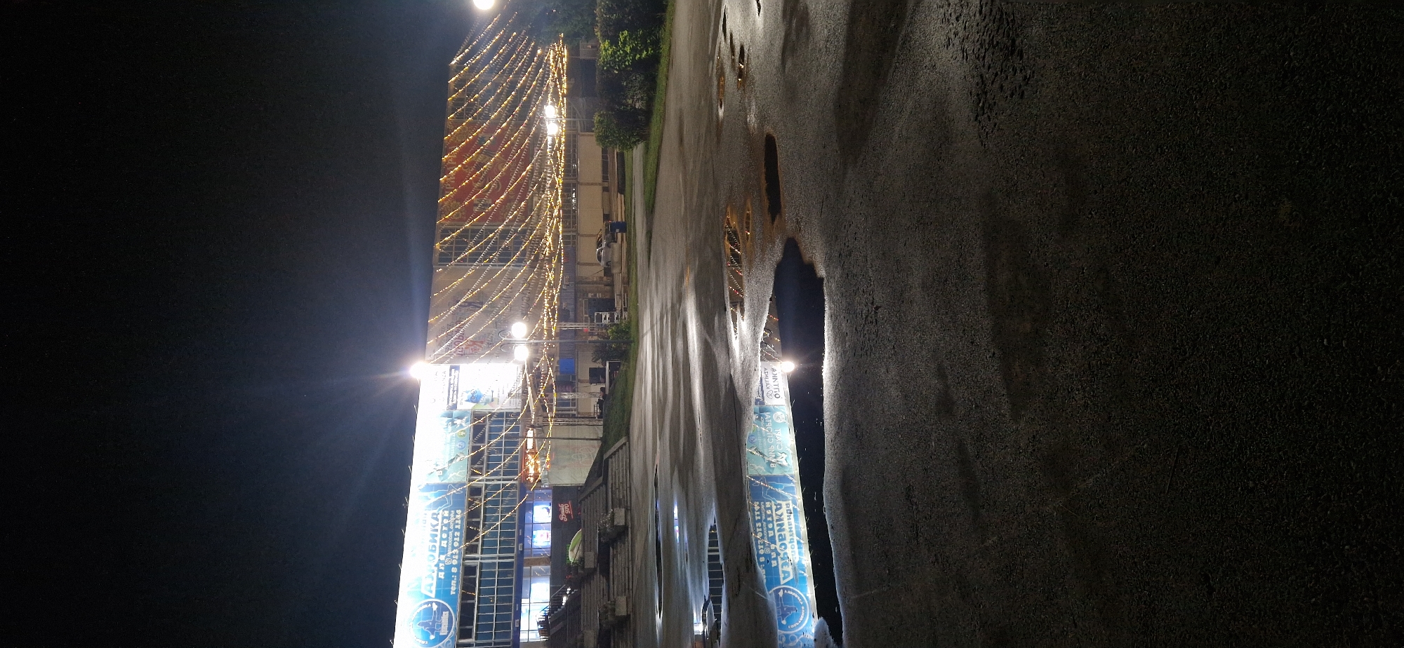 An outdoor, nighttime shot captures a modern building complex in Krasnoobsk, Russia, illuminated by strings of festive lights. The building's facade features a prominent blue and white sign with Cyrillic lettering, likely advertising a facility for children. Below this, a wide staircase leads up to the entrance. In front of the building, a wet, paved area shows several puddles reflecting the building's lights and signage. The ground appears to be asphalt, possibly wet from recent rain, with visible texture and debris. A lamppost stands near the staircase, and some trees and bushes line the right side of the scene. The sky is dark, suggesting a clear night. A few vehicles and a trash receptacle are visible in the background.