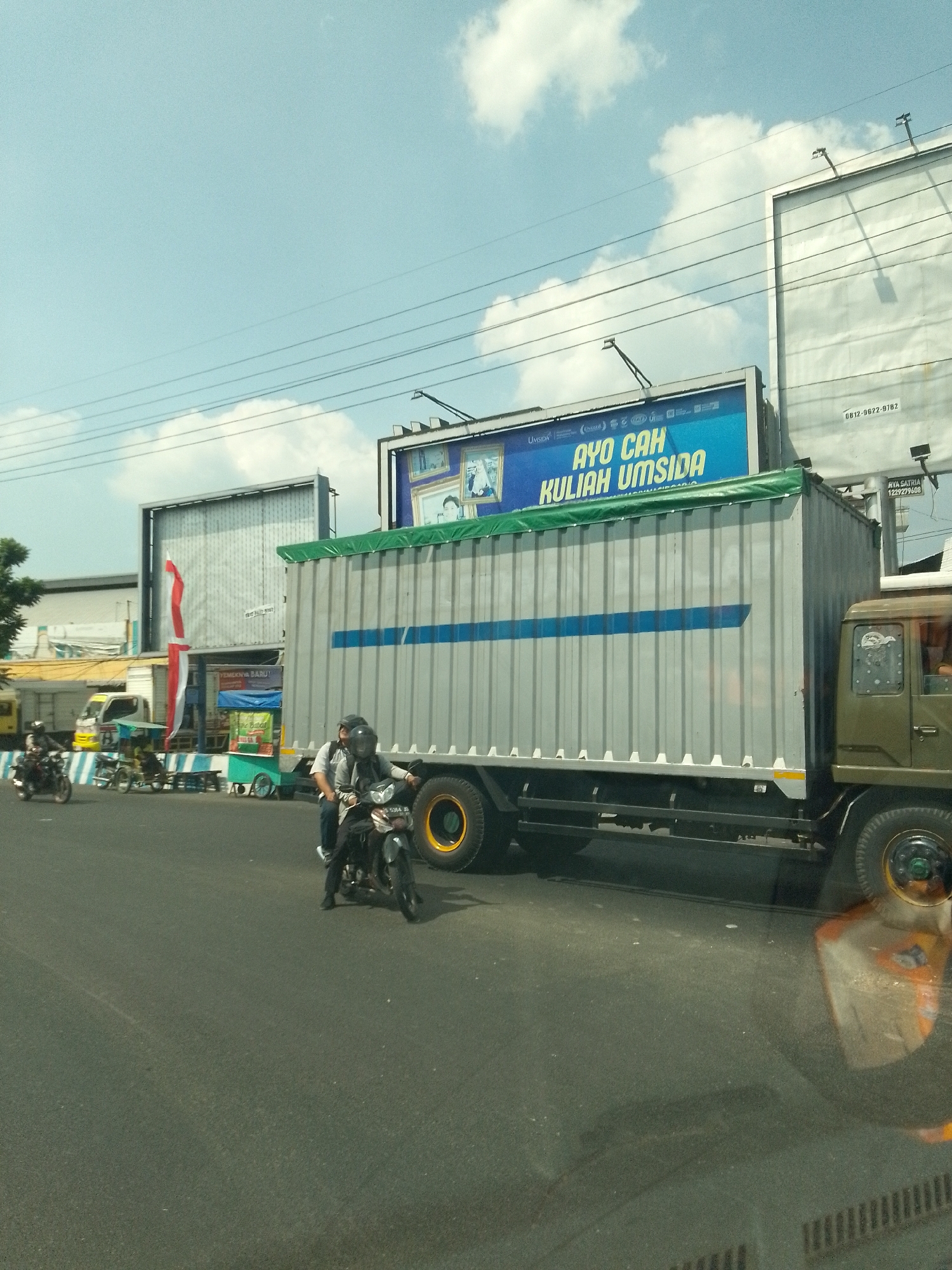 The image captures a street scene in Babat, Indonesia, under a bright, cloudy sky. A large, grey container truck with a blue stripe dominates the right side of the frame, parked on the asphalt road. Two people are riding a motorcycle in front of the truck, with the driver wearing a helmet and the passenger behind him also wearing a helmet and a grey shirt. To the left of the truck, a smaller yellow truck is visible. A billboard with a blue background and white text that reads "AYO CAH KULIAH UMSIDA" is situated above the container truck. Further to the left, another billboard with a white surface and a red and white flag attached stands in front of a building. Various other stalls and structures are present in the background, suggesting a busy commercial or urban area. The road appears to be a main thoroughfare with power lines overhead.