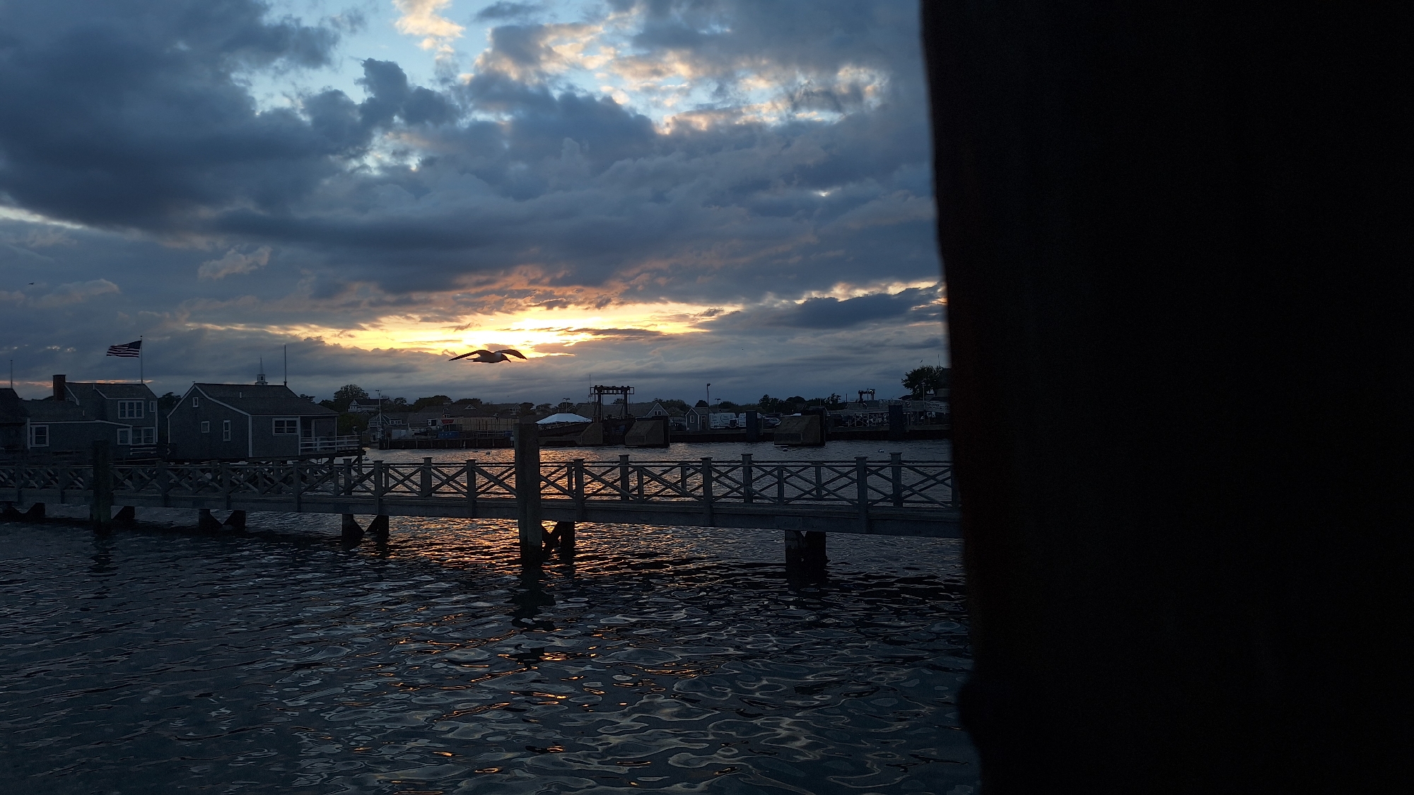 A photograph captures a tranquil sunset scene at the Nantucket harbor, Nantucket, United States of America. The sky is a dramatic canvas of dark, swirling clouds interspersed with streaks of warm orange and yellow light from the setting sun. Two seagulls are in flight, one on the left and one on the right, silhouetted against the sky. Below, the calm water of the harbor reflects the muted colors of the sky, with gentle ripples disturbing the surface. A wooden pier with a decorative railing extends across the foreground, with pilings anchoring it to the seabed. On the left side of the pier, a cluster of grey, weathered buildings with peaked roofs and American flags stand prominently. The buildings appear to be historic, possibly part of a waterfront village. Beyond the pier and buildings, the harbor stretches out, with further structures and boats faintly visible in the distance. A dark, silhouetted object, possibly a tree or a building structure, obscures the right side of the frame, adding a sense of depth and framing the view. The overall atmosphere is one of peaceful serenity, accentuated by the fading light of dusk.