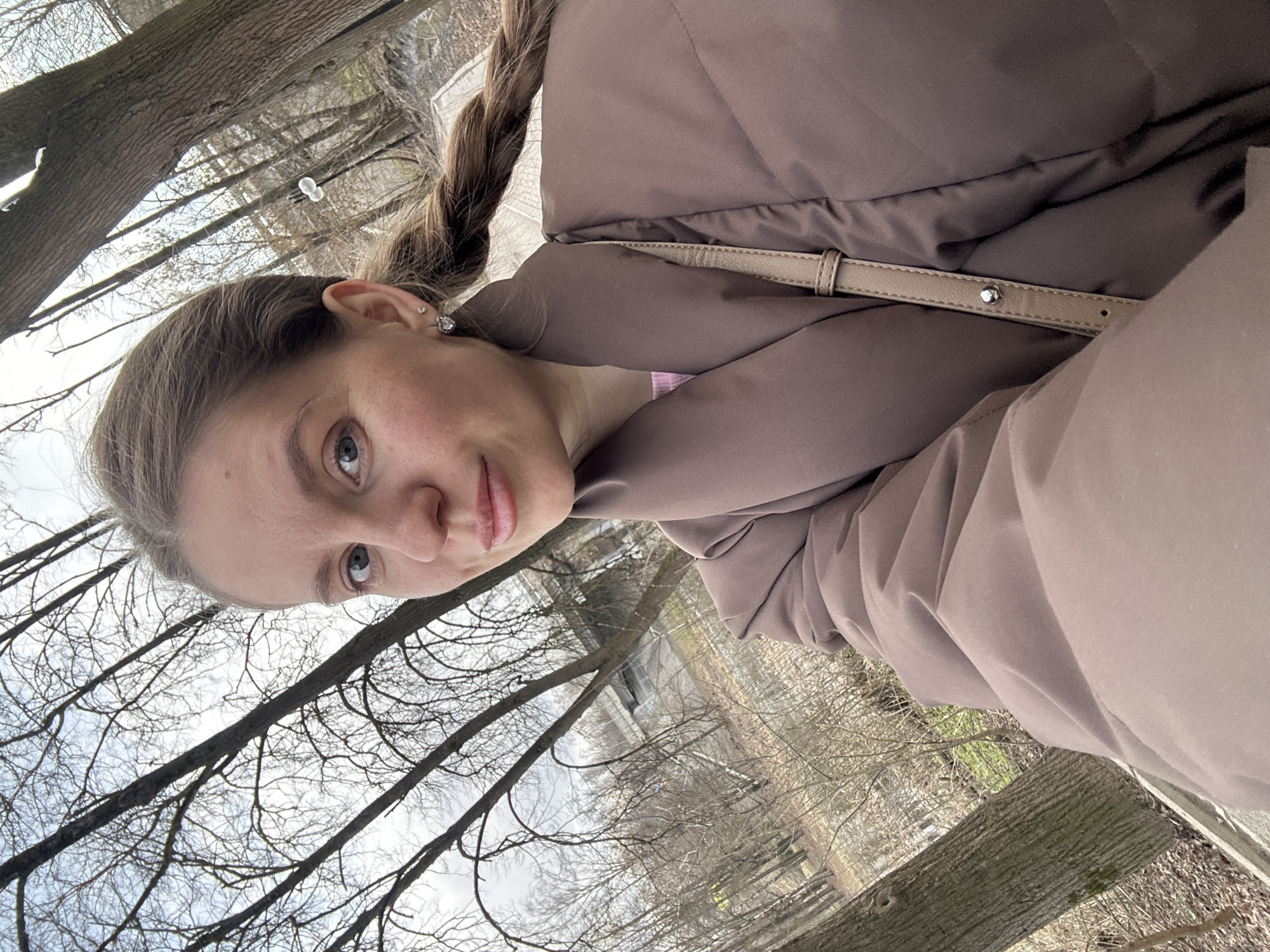 The image is a selfie of a young woman with fair skin, light brown hair braided in a partial plait, and wearing a mauve-colored puffer jacket with a light tan strap, possibly from a bag, visible over her shoulder. She is looking at the camera with a slight smile. She has small stud earrings in her ears.
The background appears to be an outdoor scene, possibly a park. Bare tree branches are visible, suggesting it is late fall or winter. In the distance, there is a bridge or some type of structure over a river. The location is most likely the city of Klin, Russia. A tree trunk is visible in the foreground.