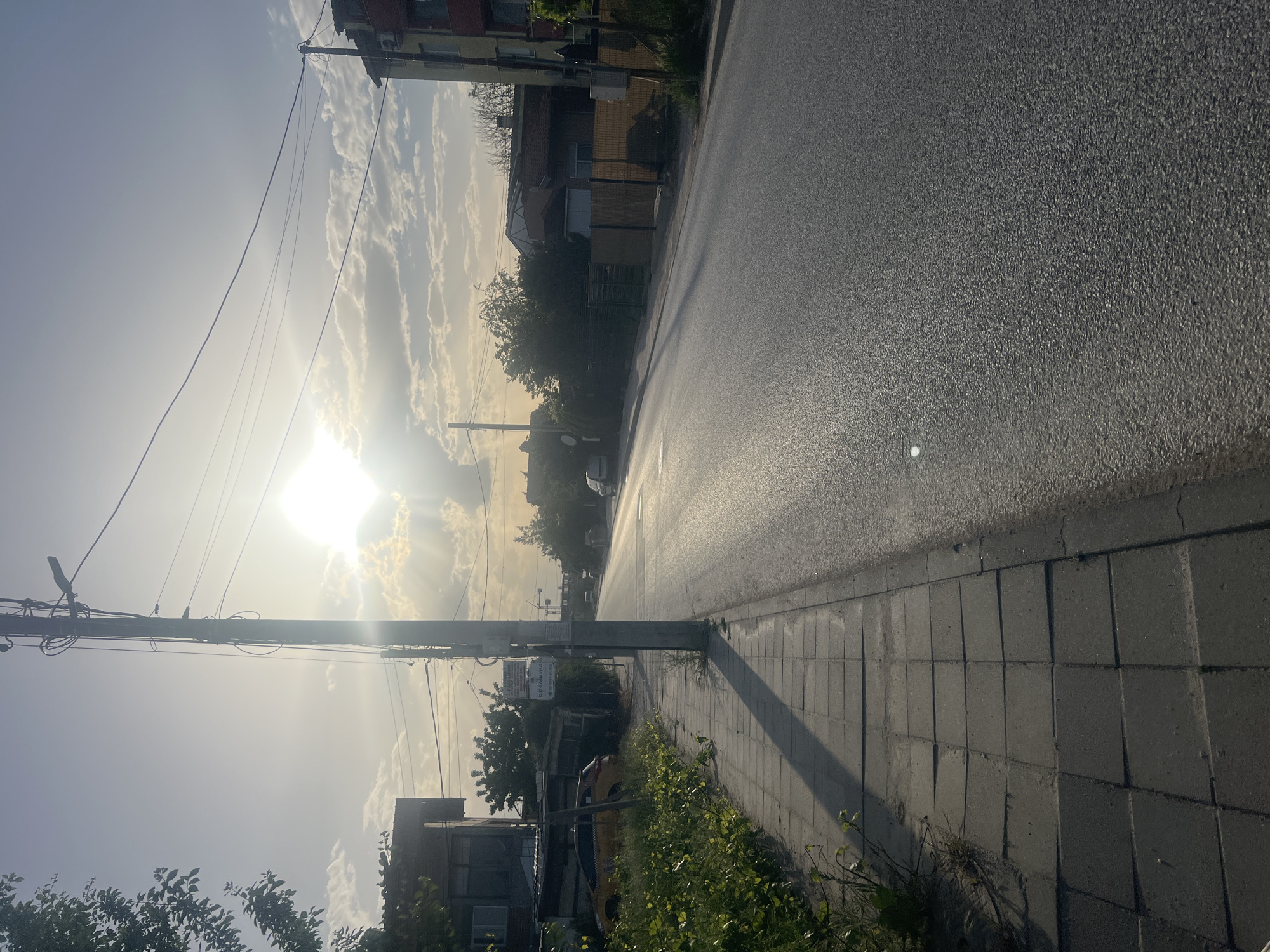 The image shows a street scene in Plovdiv, Bulgaria on a sunny day. The sun is shining brightly, partially obscured by clouds. An asphalt road is to the right, while a tiled sidewalk is to the left. Vegetation, including bushes and trees, lines the sidewalk. Buildings are visible in the background. Utility poles with wires are positioned along the left side of the street. A parked vehicle can be discerned in the distance. A sign is affixed to one of the poles.