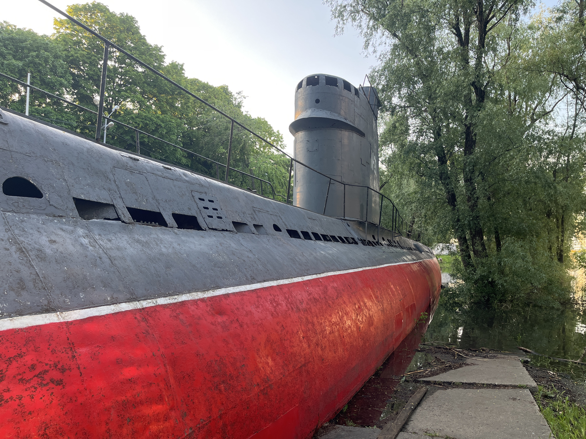 The image shows a Soviet submarine, likely a Whiskey-class submarine, exhibited as a museum piece or memorial. The submarine is painted with a dark gray upper hull and a red lower hull, separated by a thin white stripe. The conning tower is gray. A metal railing runs along the top of the hull, possibly for visitors. The submarine is situated next to a body of water, with part of the hull appearing to be submerged. Trees surround the site, suggesting a park setting. Concrete slabs form a pathway leading to the submarine.
The location is Krasnodar, Russia, based on the available information.