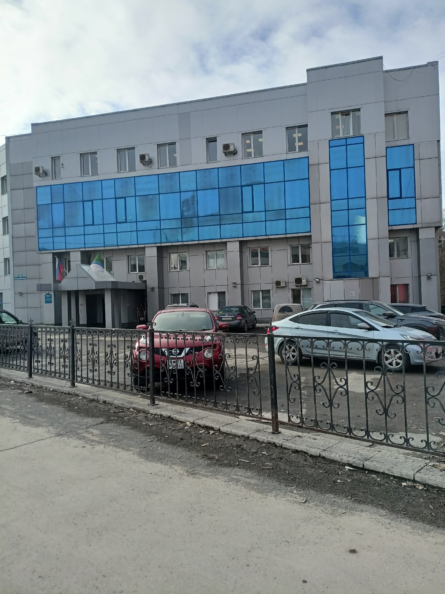 The image captures a four-story building in Novosibirsk, Russia. The building has a modern design, predominantly clad in light gray panels with a prominent band of blue-tinted windows spanning the second and third floors. Air conditioning units are visible on the building's facade, interspersed among regular windows.
In front of the building, a low decorative black metal fence separates the parking area from the sidewalk. Several cars are parked in the parking area, including a red Nissan Juke partially obscured by the fence. The Russian license plate "P 73 PC 154" is visible on the Juke. Other vehicles are parked behind it, including a light-colored sedan.
Two flags - one appearing to be the Russian flag and another, potentially the flag of Novosibirsk Oblast, are displayed above the building's entrance. A small street sign bearing Cyrillic script is mounted on the wall near the entrance.
The scene is shot during daylight hours under an overcast sky. The sidewalk is visible in the foreground, showing signs of wear.