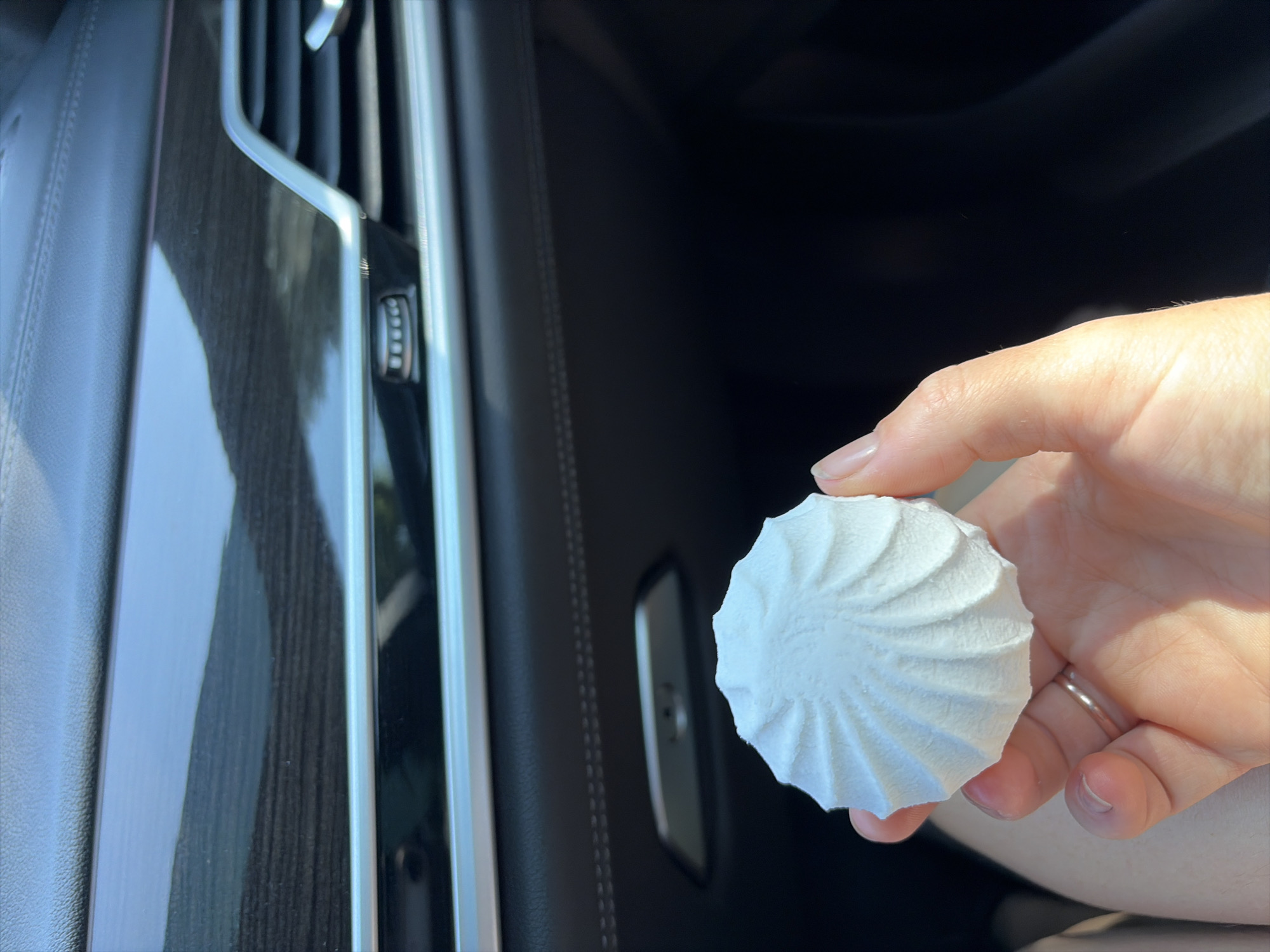 In this close-up shot taken inside a car in Criuleni, Moldova, a person's hand holds a white, swirled marshmallow. The background of the image features the dark, textured interior of the car, including a prominent wood-grain trim panel with silver accents and a section of black leather upholstery. The marshmallow, the primary subject, is positioned in the foreground, its delicate texture and spiral pattern clearly visible. The person's fingers are clasped around the sweet treat, with a silver ring visible on one finger. The lighting suggests it is daytime, with sunlight casting shadows and highlighting the textures within the car.
