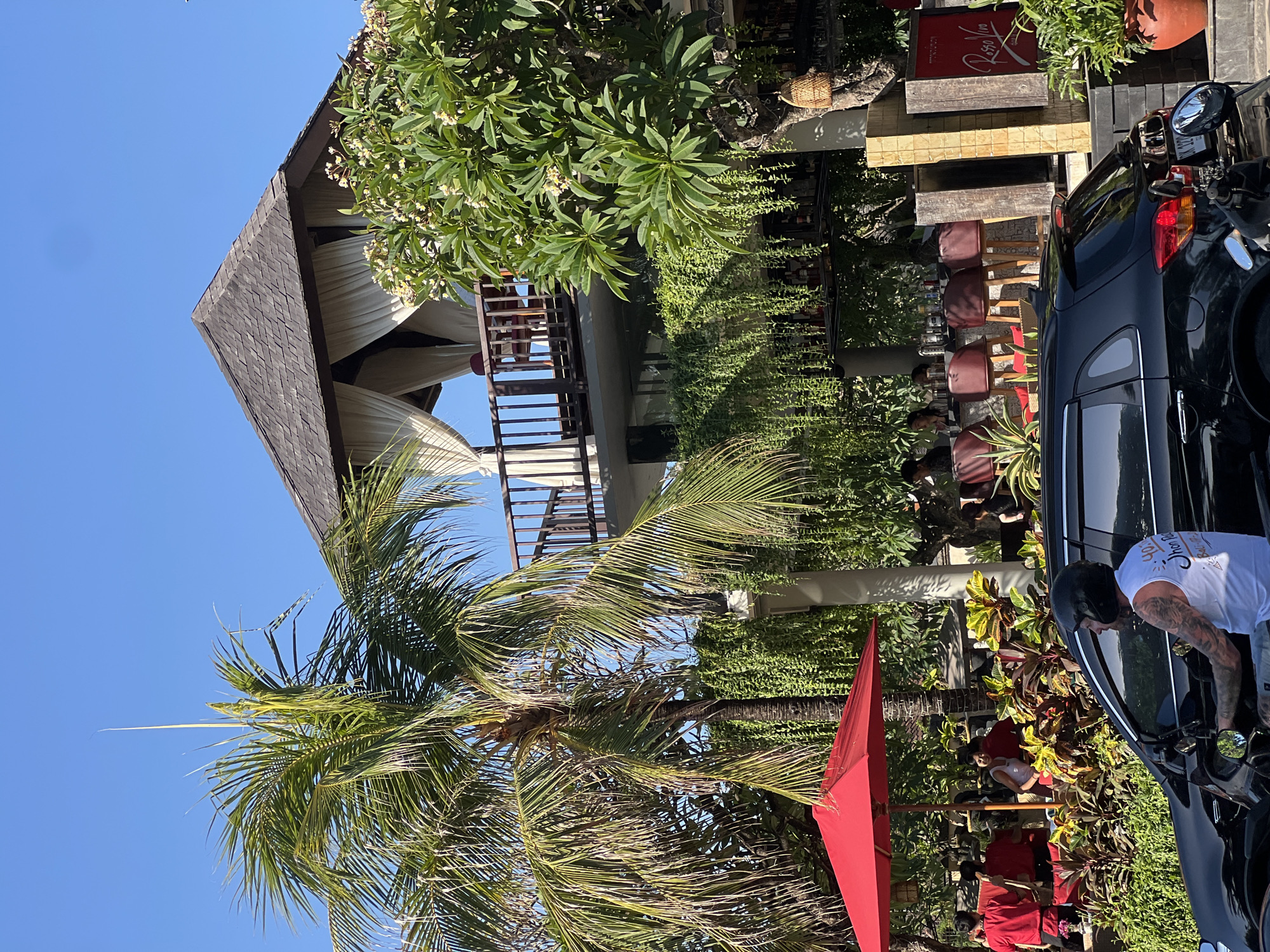 This image depicts a street scene in Kuta, Indonesia, on a sunny day with a clear blue sky. In the foreground, a black car is driving on the road, and a person is riding a motorcycle with a black helmet and a white tank top. Behind the car, a red umbrella is set up over a seating area, where several people in red shirts appear to be working. Lush greenery, including palm trees and hanging plants, dominates the middle ground, interspersed with a multi-story building featuring an open-air upper level with white drapes. The upper level has a wooden roof and railings, and tables and chairs are visible. A sign with red text reads "Rosso Vivo" on the right side of the building. The scene suggests a bustling outdoor dining or entertainment venue in a tropical location.