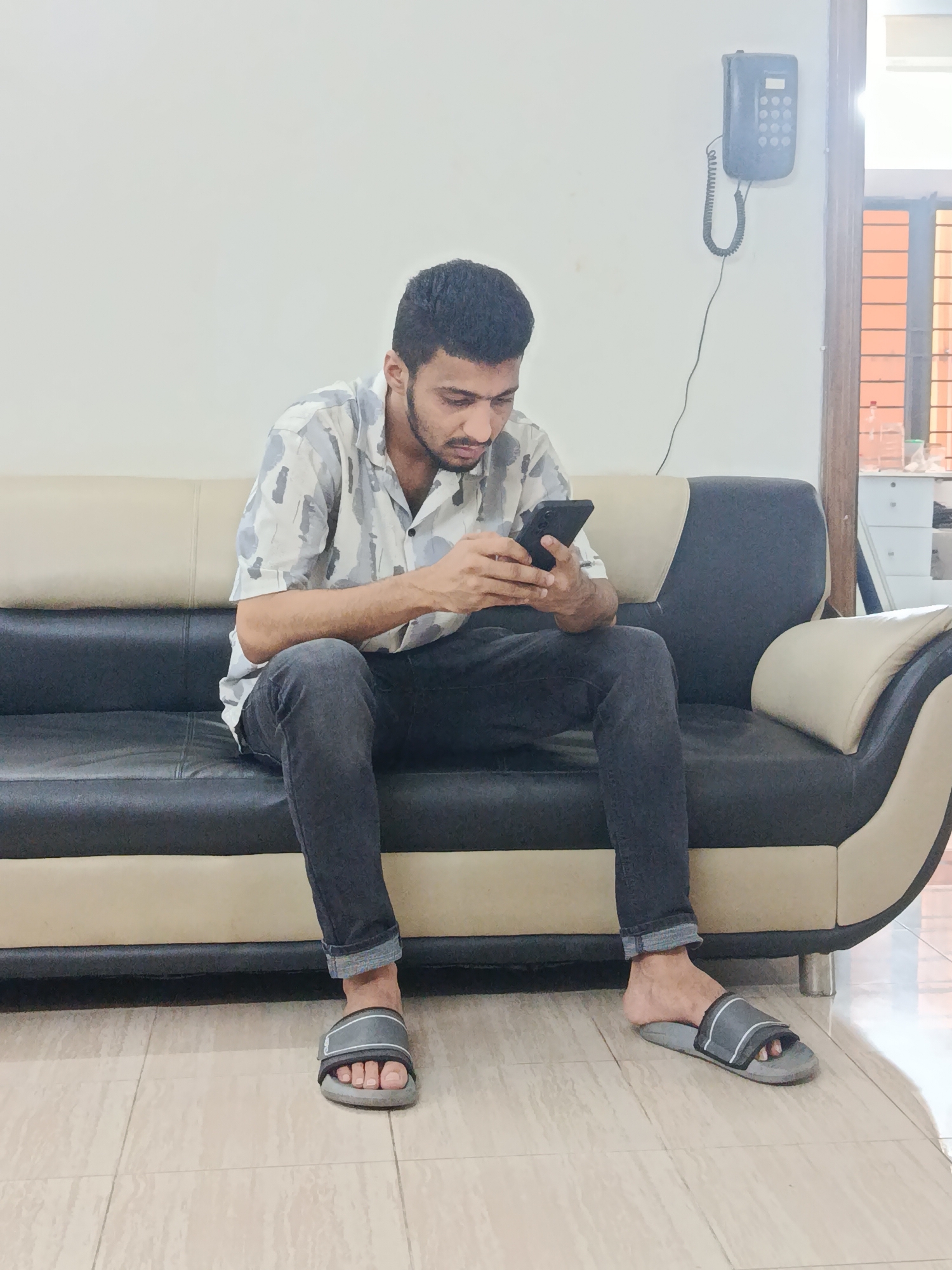 A young man is sitting on a two-toned black and beige sofa in what appears to be a domestic setting, possibly an apartment or house in Dhaka, Bangladesh. He is holding a smartphone, looking down at the screen, and appears to be engrossed in its use. He is wearing a short-sleeved shirt with a light-colored camouflage or abstract pattern, dark jeans, and gray-and-black slide sandals. A landline telephone is mounted on the wall behind the sofa. The room has a tiled floor. A window is partially visible in the background, along with a glimpse of what might be kitchen counter.