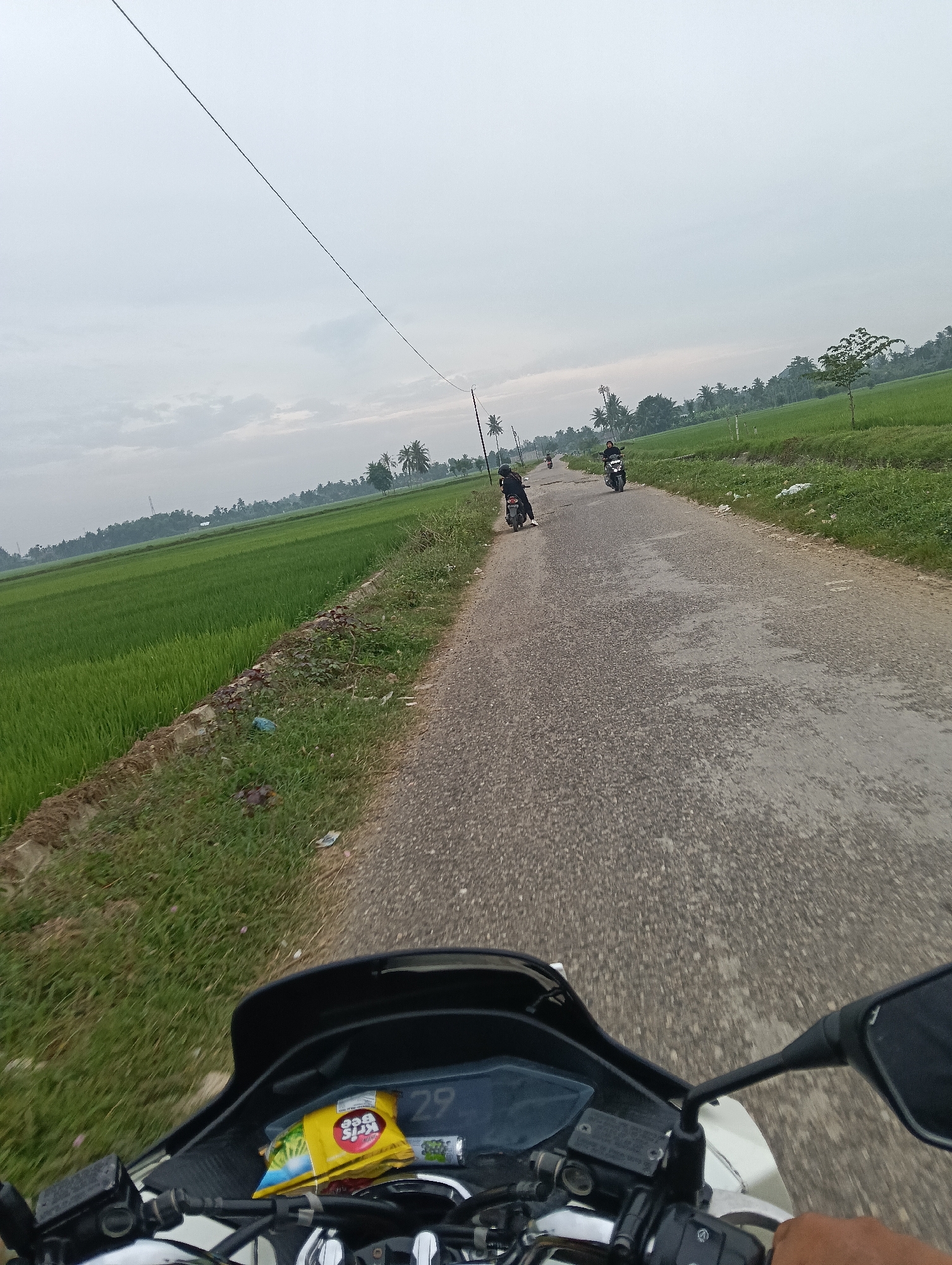 The image depicts a first-person perspective from a motorcycle traveling on a paved road in a rural area, likely Cutneuheun, Indonesia, given the presence of lush green rice paddies and tropical vegetation. The sky is overcast with a pale, diffused light. A power line stretches diagonally across the upper portion of the frame.
Several other motorcycles with riders are visible ahead on the road. One rider in black is closer to the foreground on the left, while another, also in dark clothing, is further ahead on the right. A third, less distinct motorcycle is also present in the distance.
The motorcycle in the foreground has a digital display showing the number "29". A yellow snack package, possibly containing crackers, is placed on the dashboard. The handlebars and mirrors of the motorcycle are visible, with a hand holding the right grip.
The environment consists of vibrant green rice fields bordering the road on the left, with some grass and brush along the roadside. Coconut trees and other foliage are scattered throughout the landscape, indicating a tropical setting. The road surface appears to be asphalt with some visible patches and wear.