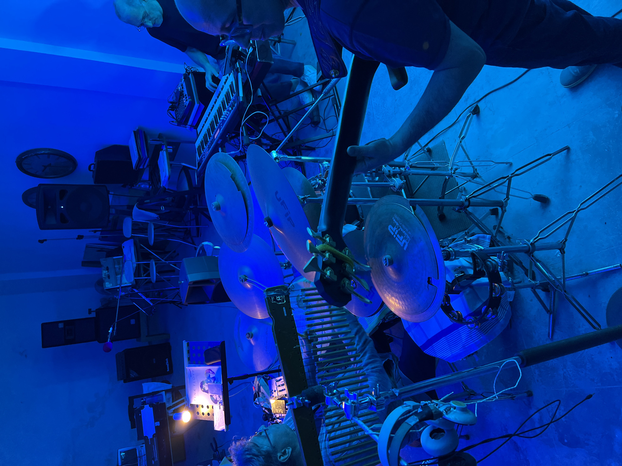The image depicts an indoor music rehearsal or recording session in Santeramo in Colle, Italy, illuminated predominantly by blue lighting. Three individuals are present, engaged with musical instruments.
On the left, a person with short, light-colored hair and glasses, wearing a striped shirt, is seated and operating a set of chime bars or wind chimes with both hands. A music stand in front of them holds two album covers; the top one is clearly identifiable as Pink Floyd's "The Dark Side of the Moon," illuminated by a bright, bare light bulb. White over-ear headphones hang from a stand in the bottom left foreground.
In the right foreground, a person wearing a dark short-sleeved shirt and dark pants holds and plays an acoustic guitar. Their right arm is positioned over the guitar body. Visible on the floor are various cables and the person's light-colored shoes. Adjacent to this individual, multiple cymbals, including one branded "UFIP," are mounted on stands, along with a tambourine.
In the right background, another person with short, light-colored hair and glasses, dressed in a dark short-sleeved shirt, operates a keyboard synthesizer with both hands. Next to the keyboard, a smaller electronic device with knobs and buttons is visible.
The room's walls are painted a light color, appearing intensely blue due to the stage lighting. The floor is a light-colored concrete or industrial surface. Various audio equipment is distributed throughout the space, including several large speakers, some marked "SoundLab," positioned on elevated surfaces or stands. Microphone stands are also present. In the background, two large, circular wall clocks are mounted on a wall. The overall environment suggests a functional music studio or rehearsal space, equipped with professional instruments and audio gear.