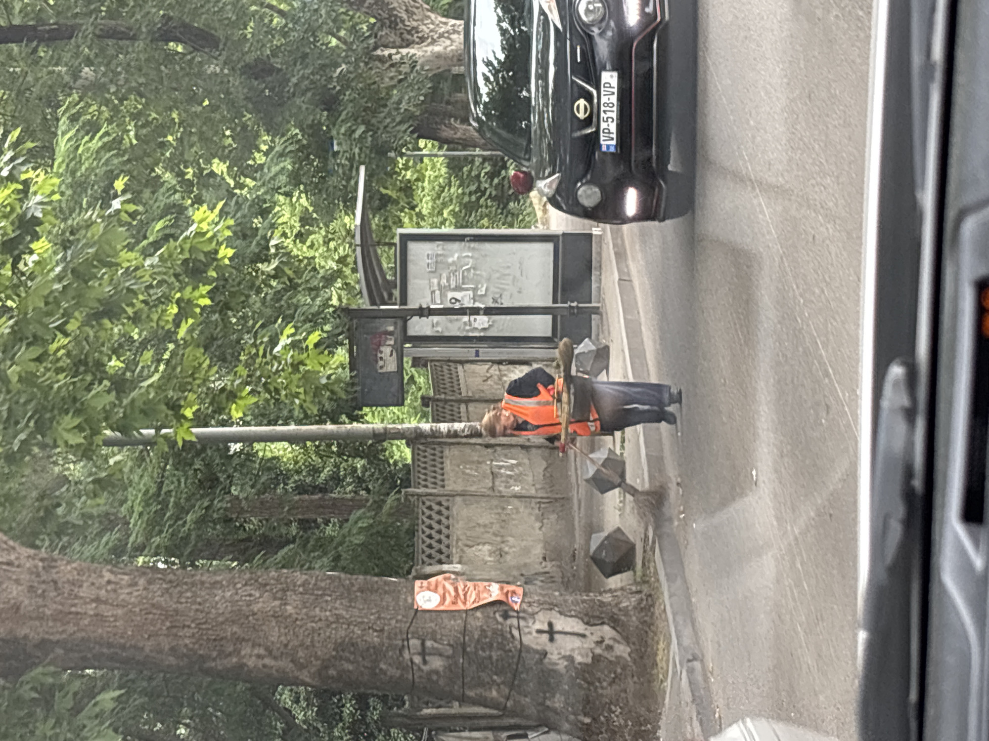 A woman in an orange safety vest is sweeping the sidewalk with a broom near a bus stop. She is wearing a dark jacket and pants. She is holding a dustpan in her other hand. There is a large tree to her left with two crosses painted on it, one with an orange banner draped over it. In the background, there is a gray fence and a bus stop shelter. A black car with a license plate that reads "VP-518-VP" is driving on the road to the right of the woman. The scene appears to be in an urban environment.