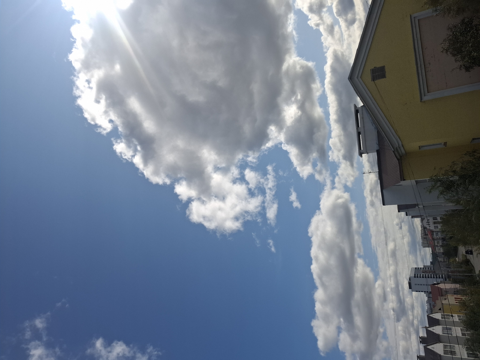 The image depicts a view of a residential area in Yakutsk, Russia, under a partly cloudy sky. In the foreground, the roofline of a yellow building with a white trim and a ventilation grate is visible. Several other buildings, including some with steeply pitched roofs, are seen in the middle ground, suggesting a neighborhood of houses. In the distance, a tall, modern building stands out against the horizon. The sky is dominated by a large, fluffy cloud formation illuminated by sunlight, with patches of blue sky visible. Power lines stretch across the scene, indicating urban infrastructure. A single car is visible on a road winding through the neighborhood. The overall impression is a daytime scene in an urban or suburban setting, characterized by a mix of architectural styles and the natural element of clouds.