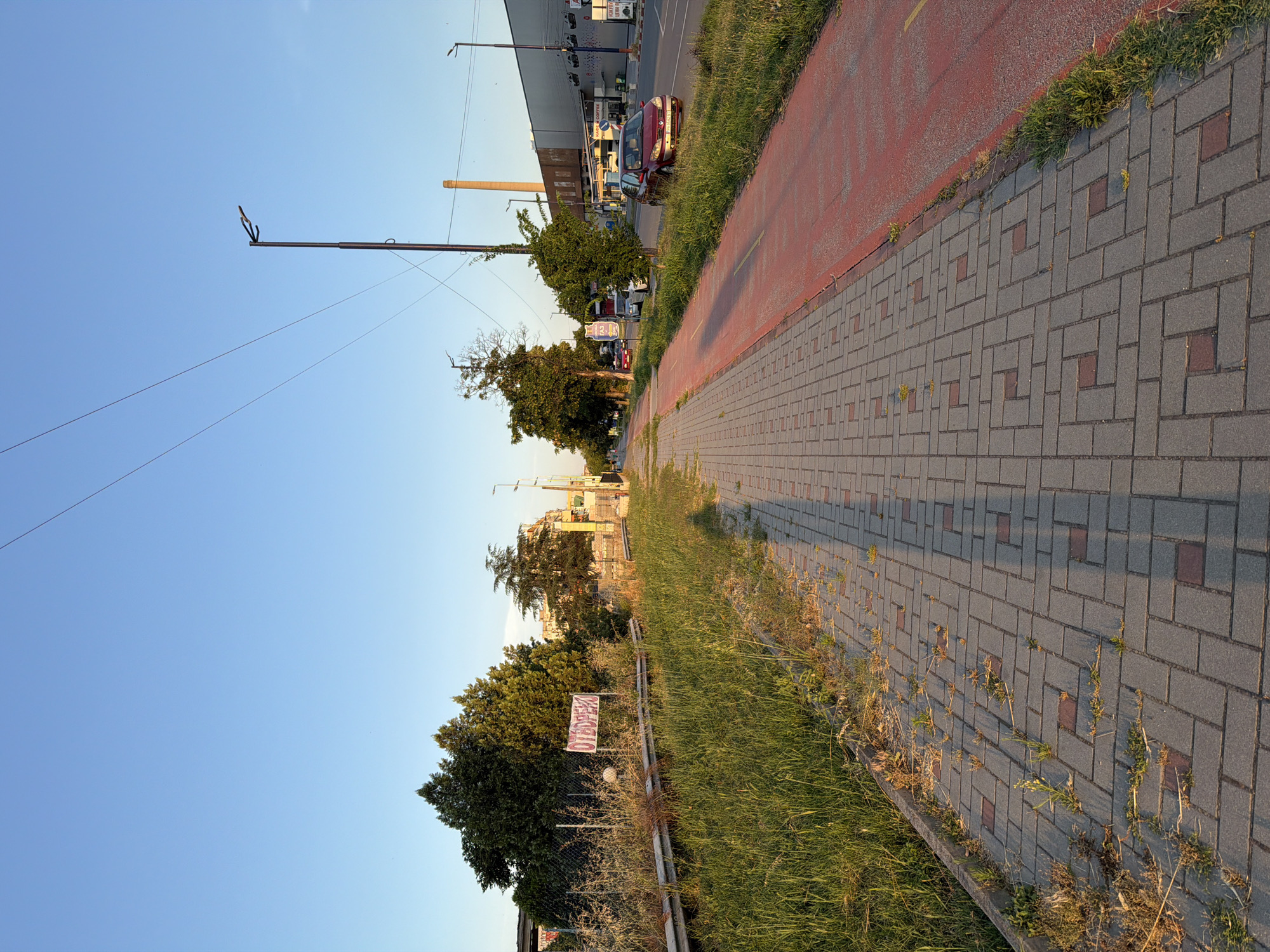 This is a street-level, vertical photograph taken in Pazardzhik, Bulgaria, showing a paved pedestrian walkway and a dedicated red-painted bicycle lane alongside a grassy verge and a road with light traffic. The scene appears to be captured during the late afternoon or early evening, as indicated by the long shadows cast on the walkway.
On the left side of the image, a grassy area is bordered by a metal guardrail and a chain-link fence. Behind the fence, several trees are visible, with a banner displaying the word "ОТВОРЕНО" (meaning "Open" in Bulgarian) in pink capital letters hanging from a post. Further back, more trees and some buildings can be seen.
The walkway itself is paved with gray interlocking bricks arranged in a geometric pattern. Weeds are growing in some of the gaps between the bricks. A prominent shadow of a person, presumably the photographer, stretches across the walkway from left to right.
To the right of the walkway is a red-painted bicycle lane, marked with a yellow line. Beyond the bicycle lane is a grassy area, and then a road where several cars are driving or parked. The most prominent vehicle is a red sedan on the right side of the road. In the background, a tall industrial chimney and a large, modern building are visible, suggesting an area that is likely a commercial or industrial zone. The sky above is clear and blue, with power lines crossing the frame. The overall impression is one of an urban edge, where pedestrian and cycling infrastructure meets more developed commercial or industrial areas.