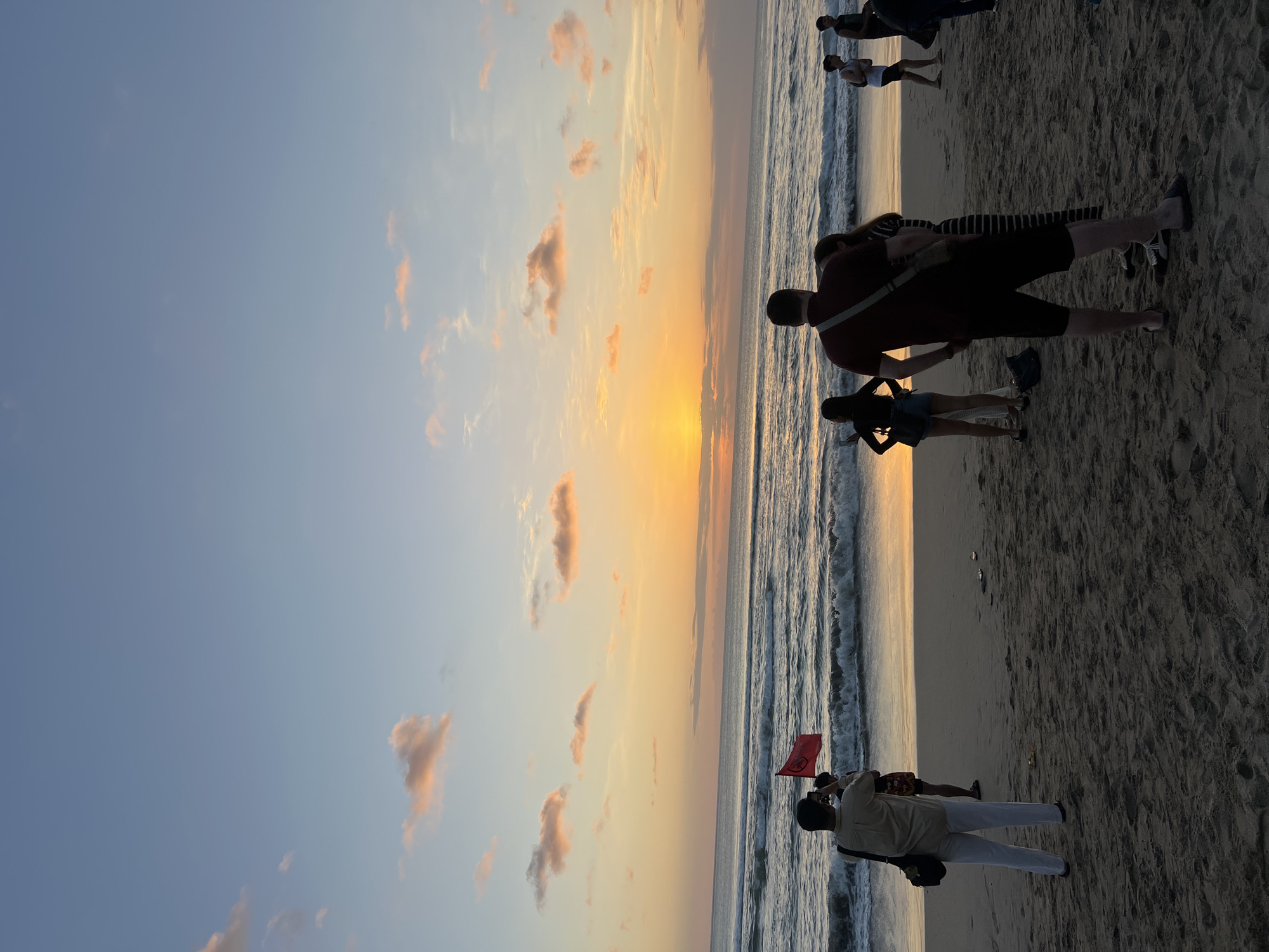 The image captures a sunset scene on Kuta Beach, Bali, Indonesia. The sky is painted with hues of orange, yellow, and soft pink, with scattered clouds. The sun is setting over the ocean, casting a warm glow on the water and the wet sand. Gentle waves are rolling onto the shore.
On the beach, several people are visible, mostly silhouetted against the sunset. In the foreground, a man in a light-colored shirt and white pants stands facing the ocean, holding a small red flag. To his left, another person is taking a photo with a phone. Further back, a group of people are gathered, including a man in a dark red shirt and shorts, and a woman in a striped dress. Other individuals are walking along the beach or standing and observing the sunset. The sand is textured with footprints and some small debris. The overall atmosphere is peaceful and serene, indicative of a popular tourist spot during sunset.