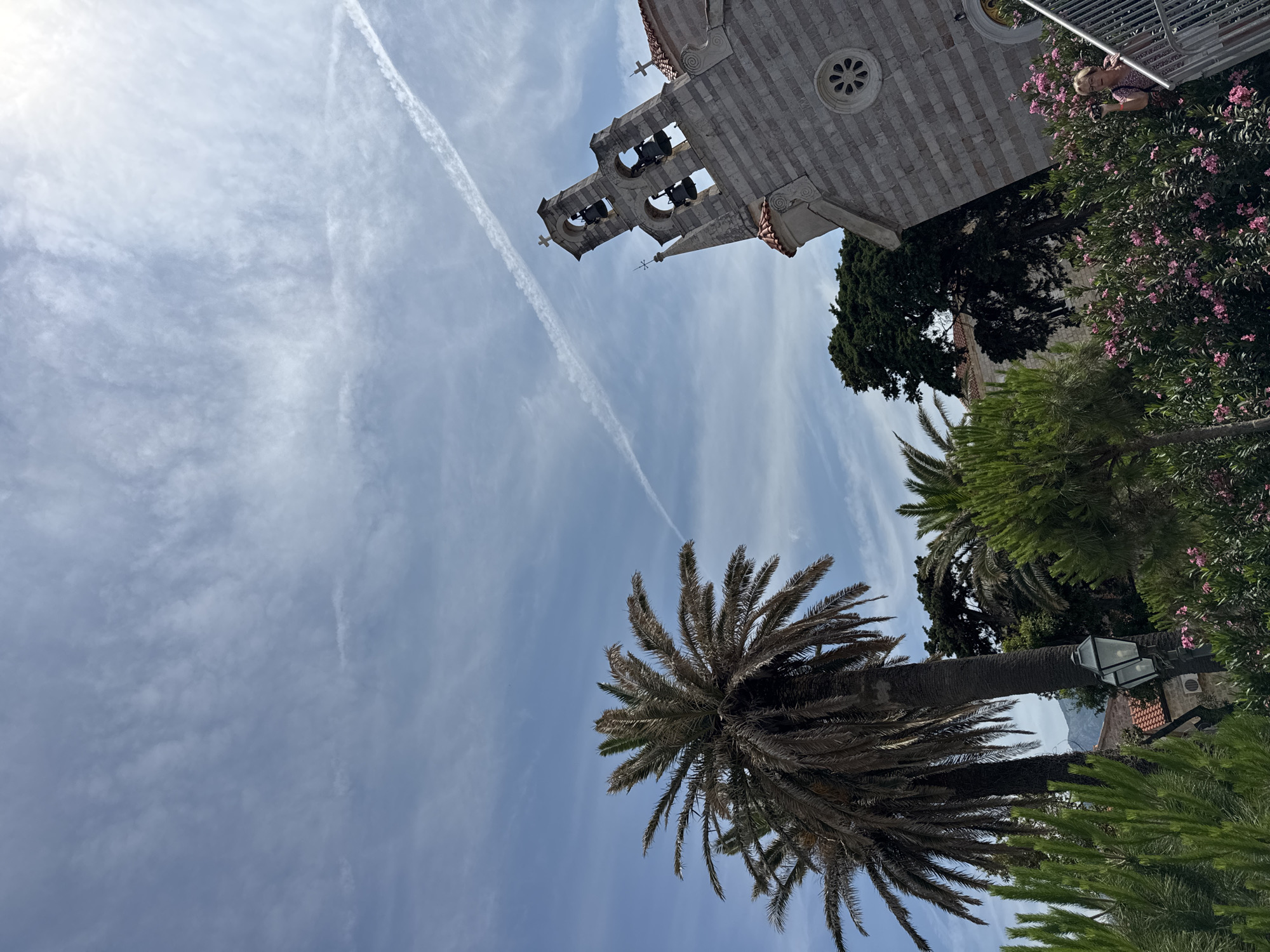 The image captures a view of a stone church with a bell tower against a partly cloudy blue sky with a prominent contrail. In the foreground, there are several palm trees and dense greenery with pink flowers. To the right, a woman is visible on a staircase, holding a camera. The architectural style of the church, particularly the bell tower and facade, suggests it could be the Church of St. Sava in Budva, Montenegro, a known landmark.