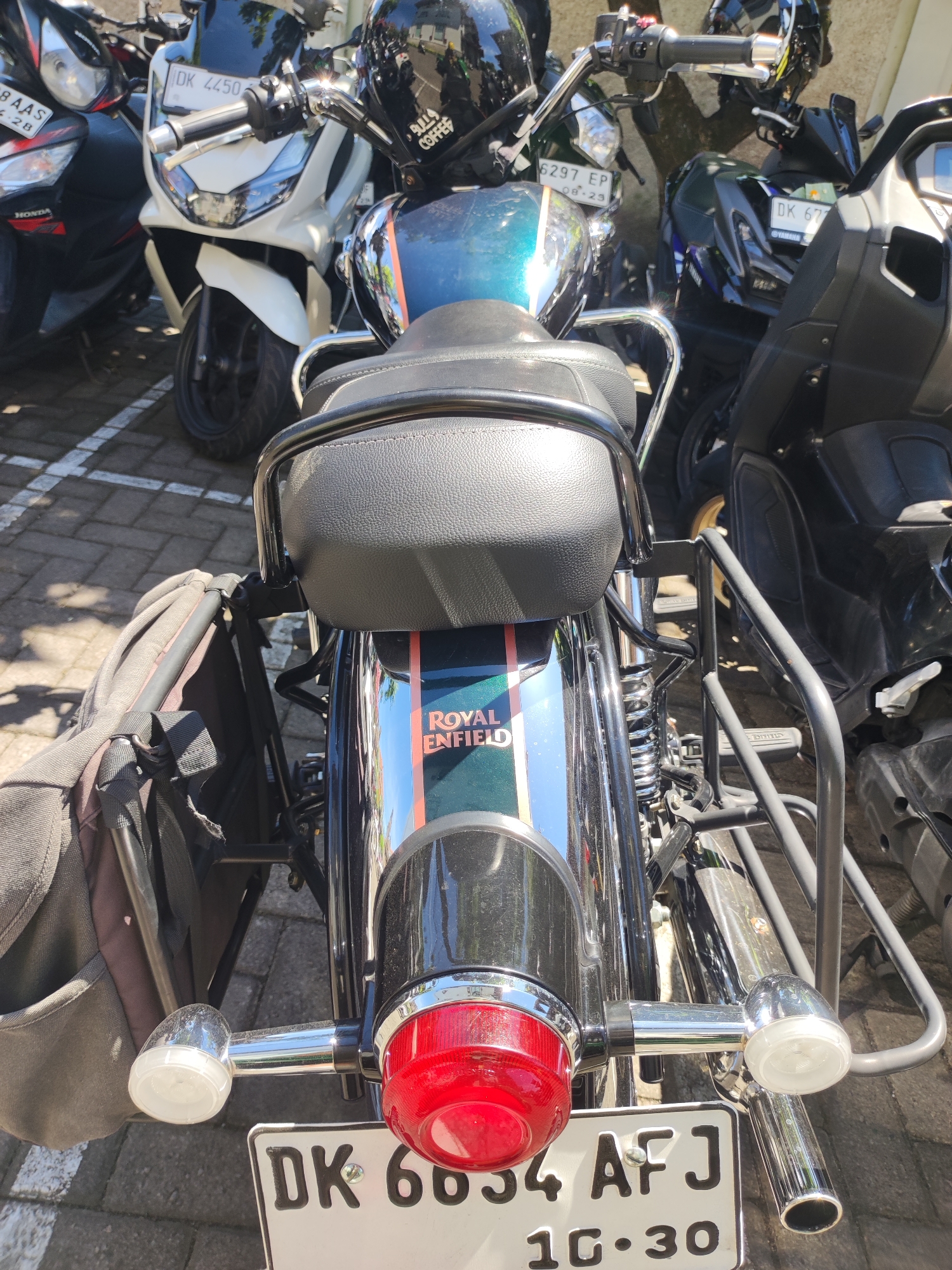 The image shows the rear of a parked Royal Enfield motorcycle with a blue and chrome finish, identifiable by the "ROYAL ENFIELD" logo. It has a black leather seat and luggage racks. A black backpack is attached to one side. The license plate reads "DK 6634 AFJ" and "10.30." The motorcycle is surrounded by other parked motorcycles and scooters, including a Honda and a Yamaha. Other license plates such as "DK 4450," "6297 EP," and "DK 677" are visible. The scene appears to be in Denpasar, Indonesia, based on the "DK" license plate prefix commonly used in Bali.