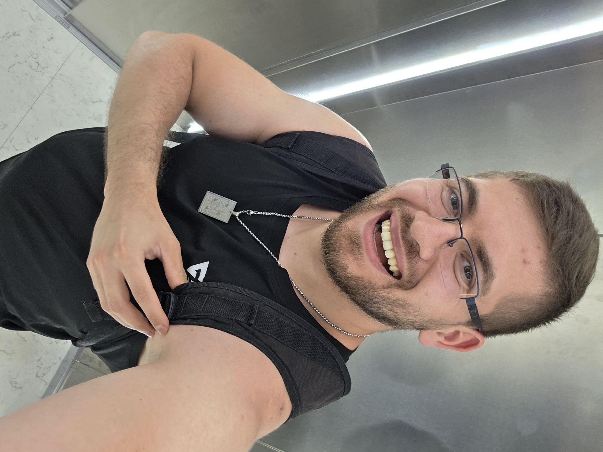 A man wearing glasses and a black tank top is taking a selfie. He is smiling and appears to be indoors, possibly in an elevator, given the stainless steel walls and ceiling with fluorescent lighting. He is wearing a silver chain necklace with a pendant. The location cannot be confirmed as Plovdiv, Bulgaria from the image. The background is blurry.