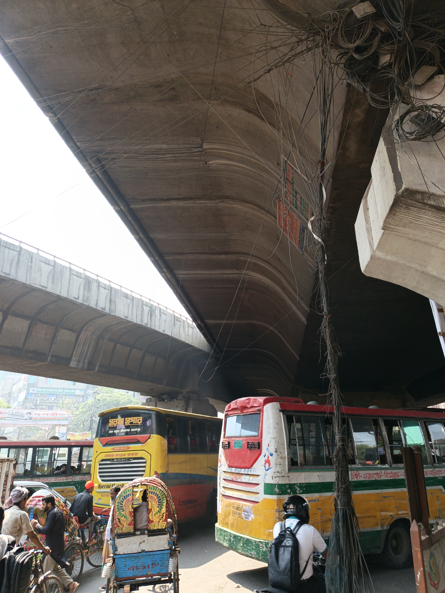 The image captures a street scene in Dhaka, Bangladesh, under an overpass. Two buses, one predominantly yellow and the other red and yellow, are prominent in the foreground, along with people on bicycles and rickshaws. A motorcyclist with a backpack is stopped behind the yellow bus. A dense cluster of electrical wires hangs from the overpass, creating a chaotic visual element above the buses. The overall scene suggests a busy transportation hub in an urban environment.