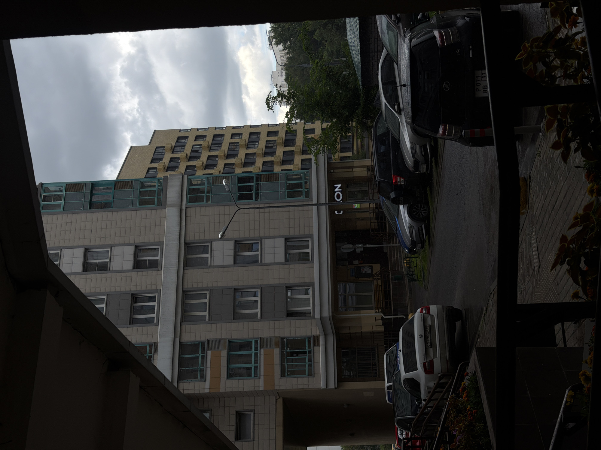 The image shows a street-level view of a residential area with apartment buildings and parked cars under a cloudy sky. The foreground features a dark, covered walkway or balcony with plants visible below. Beyond this, a light-colored apartment building with numerous windows and balconies is prominent. To the right of this building, a taller, yellowish apartment building with a brick facade stands. Several cars are parked on the wet asphalt of a street or parking area. The license plates on some of the cars are partially visible. One car in the foreground has a license plate that appears to read "C529CC 190". Another car towards the right has a license plate that reads "P 071 BB 97". The presence of these license plates suggests the location is likely in Russia. The architecture of the buildings and the visible signage on the lower level of the light-colored building, which includes the word "ICON" in white lettering, are consistent with modern urban development in Moscow, Russia. The overall atmosphere is somewhat overcast and possibly after rain, given the wet pavement. There are no people visible in the image.