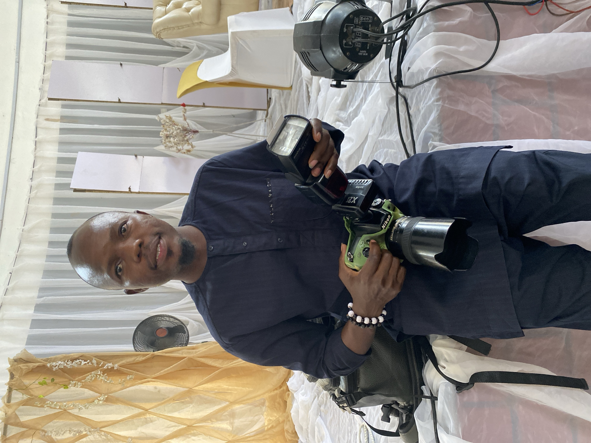 A Nigerian man, appearing to be in his late 20s to early 30s, is seated in a well-lit indoor setting that appears to be a wedding or event venue in Bukuru, Nigeria. He is wearing a dark blue traditional Nigerian outfit. The man is holding a green Nikon camera with a lens attached in his right hand and a camera flash in his left hand. He is looking directly at the camera with a neutral expression. In the background, white and gold drapery are visible, along with a floor fan, a stage light, and chairs. The scene is set on a raised platform covered with white fabric.