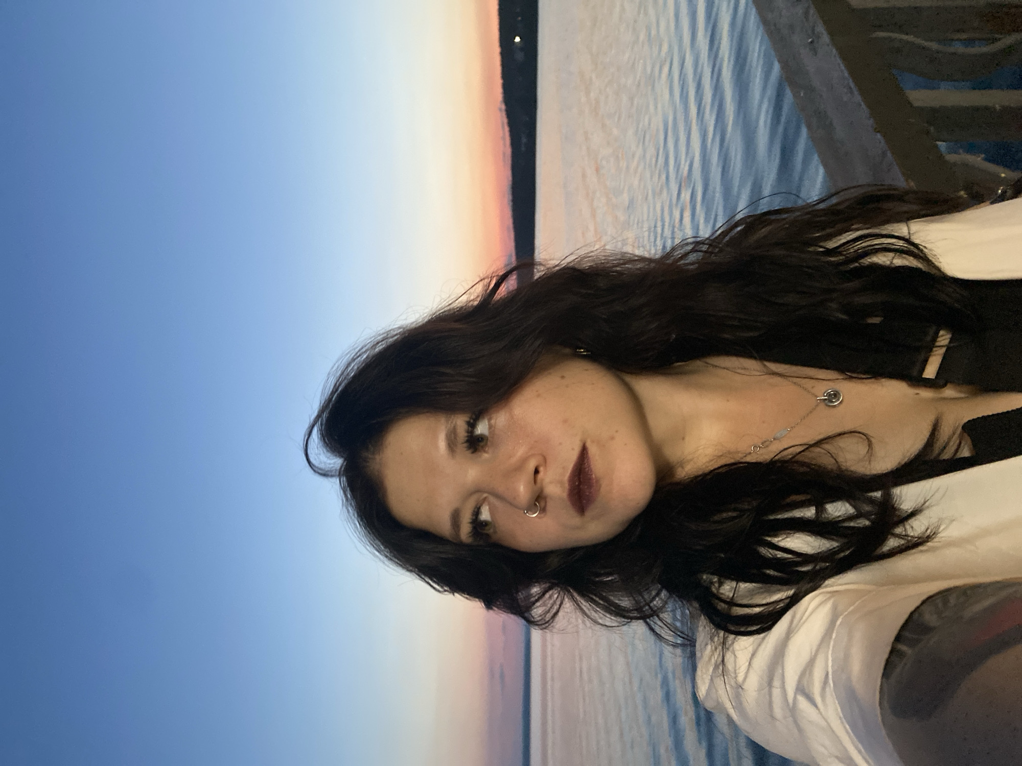 A woman with dark, wavy hair and a nose ring poses for a selfie outdoors at sunset. She is wearing a white blazer over a dark top and a silver necklace. The background shows a body of water with a faint, pink and orange sunset sky above it. A dark land mass is visible in the distance across the water. The scene appears to be on a pier or boardwalk, with a railing visible in the lower right corner.