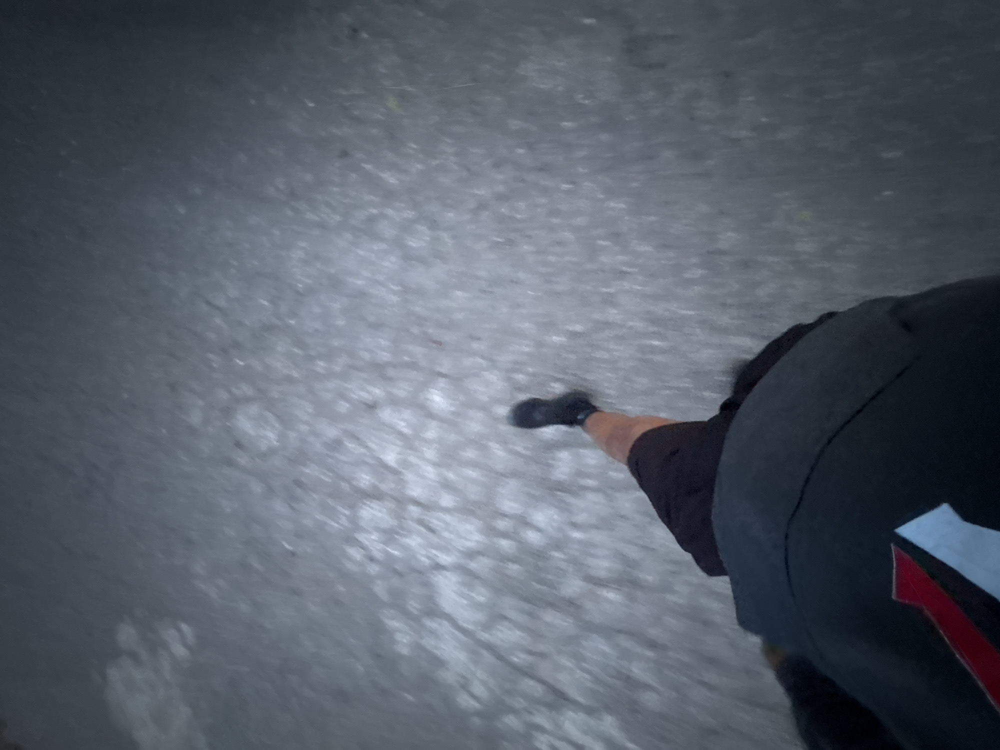 The image shows a person walking on what appears to be a paved surface. The person is wearing black shoes, black shorts, and a black top with a red and white design. The ground appears to be a gray or light-colored paved surface. The image is taken from a high angle, looking down at the person's legs and feet. The location is not specified.