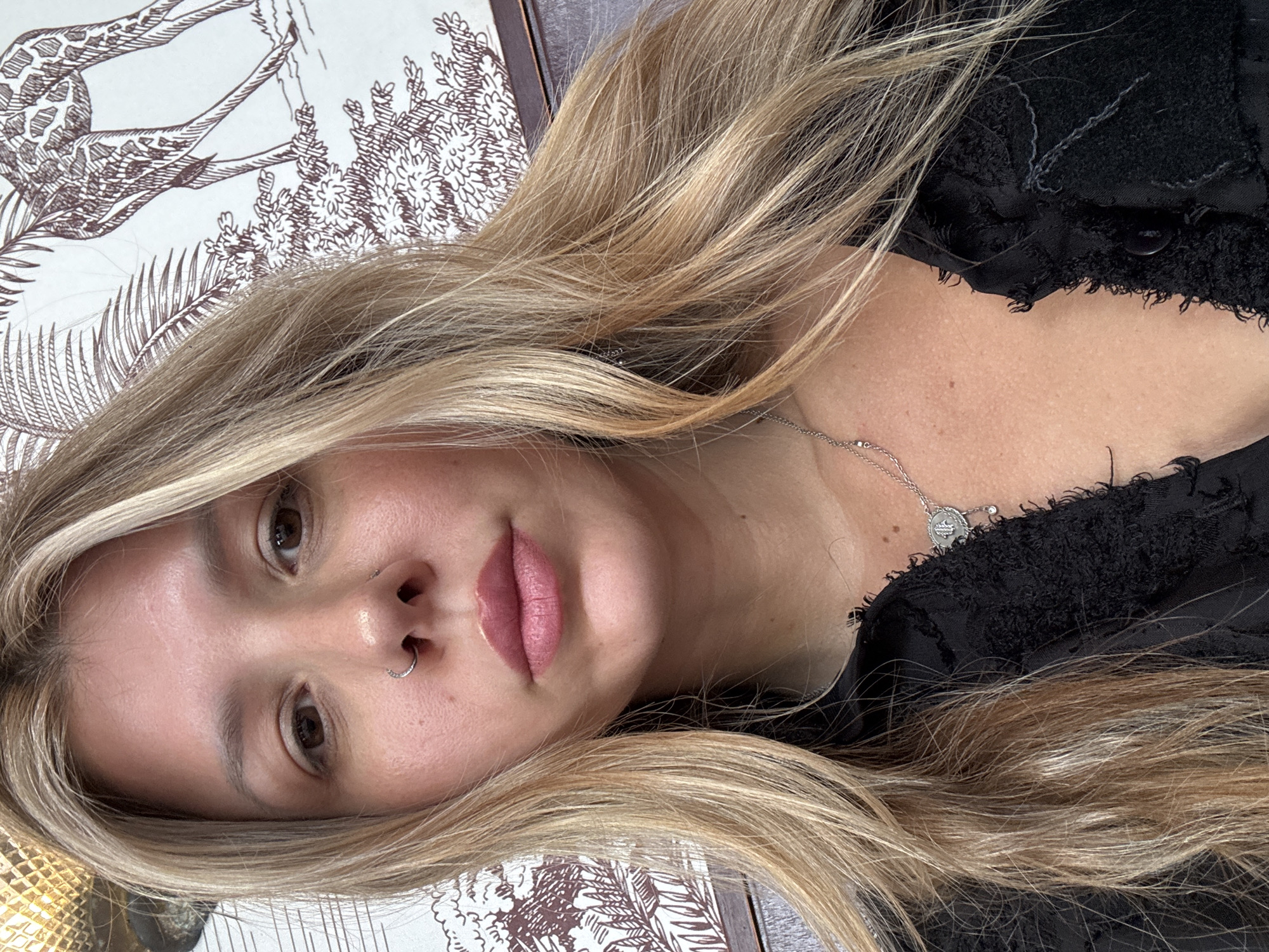 A close-up, head-and-shoulders selfie shows a young Caucasian woman with blonde hair and a nose ring. She is wearing a black, distressed-fabric top with a silver pendant necklace. The background features a patterned wallpaper with drawings of giraffes and foliage. The lighting is natural and casts soft shadows. The image was taken in an indoor setting.