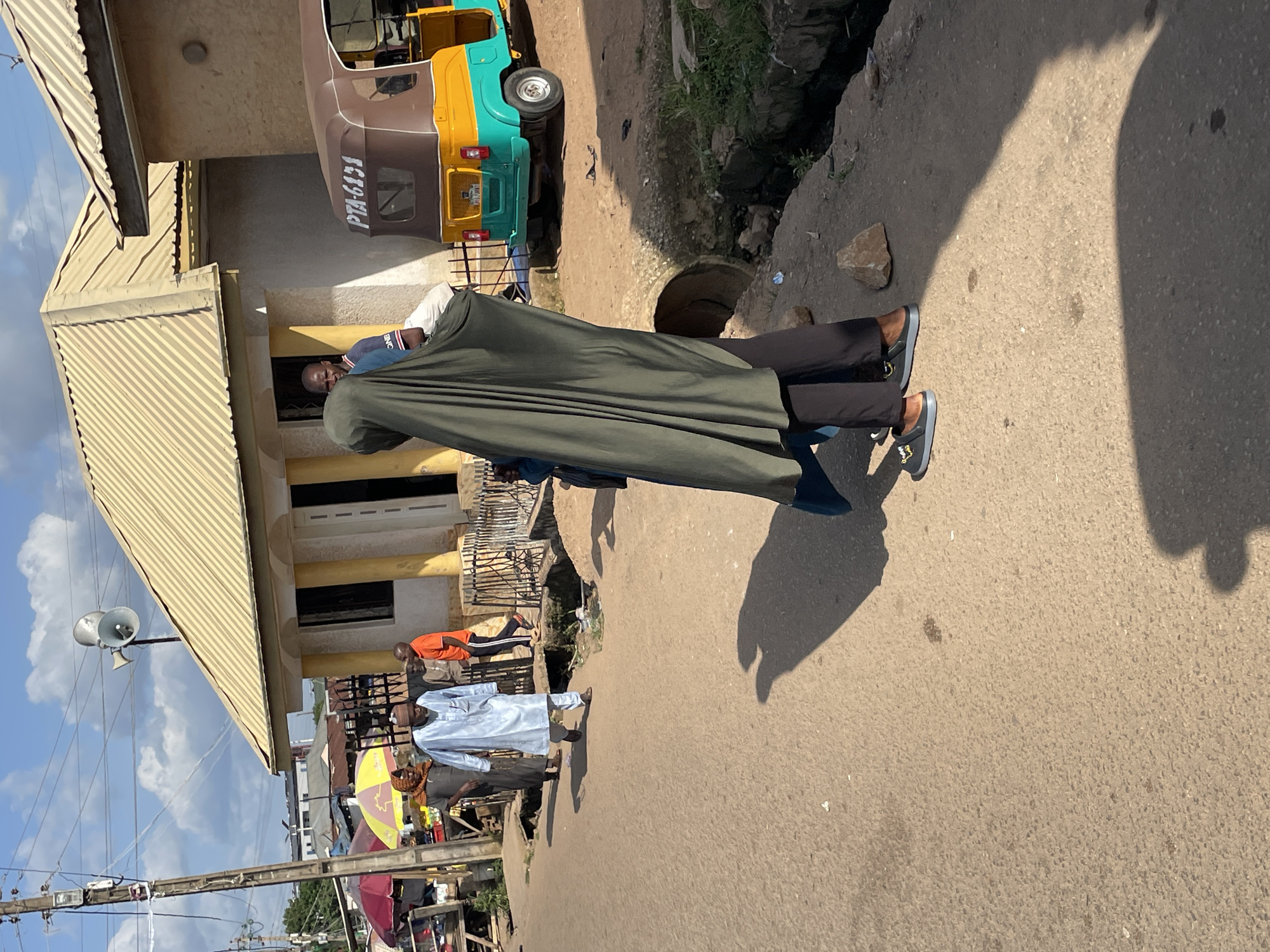 In Gura Top, Nigeria, the image captures a street scene under a bright, cloudy sky. In the foreground, a person wearing a dark green hijab and black pants walks away from the camera. They are wearing dark shoes with a yellow design on them. To their left, a man in a light blue robe and matching pants, also wearing a cap, walks towards the camera. Behind him, another man in a dark garment is visible. Further back, a man in an orange t-shirt and black pants walks towards the left of the frame. The background features a yellow-roofed building with white trim and columns, and a brown and turquoise motorized tricycle with the license plate "PTA-6434". Various market stalls with umbrellas are present on the left side of the image. The street is paved with asphalt, and a drainage channel runs along the right side. Electrical poles and wires are visible overhead.