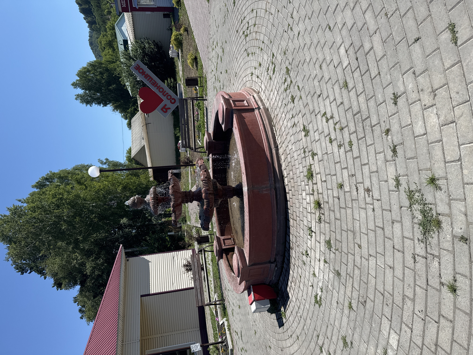 The image depicts an outdoor scene with a fountain as the central focus, situated on a paved circular area. The fountain is made of a dark reddish-brown material and has multiple tiers with water cascading down. It is surrounded by a circular red basin. To the left of the fountain, there is a light-colored building with a red roof and rolling shutters. A wooden bench is visible in front of this building. In the background, trees and some low-rise buildings are present. A prominent sign with a red heart and the text "Я СОЛОНЕШНОЕ" (I love Soloneshnoye) is visible to the right of the fountain, angled upwards. The ground is paved with grey interlocking bricks, and some grass is growing in the gaps. The sky is clear and blue. The overall environment suggests a public space in a town or settlement, likely Soloneshnoye, Russia, given the signage. There are no people visible in the image.