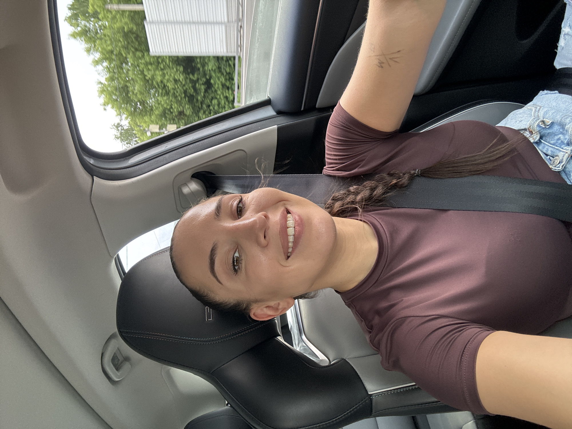A woman with a braided ponytail is taking a selfie while driving a car. She is wearing a maroon t-shirt and denim shorts, and her seatbelt is fastened. The interior of the car features black and gray leather seats. Through the car window, a blurry background of green trees and a white barrier is visible, suggesting an outdoor environment possibly along a highway or road. The woman's right arm has a tattoo or marking. The scene takes place during the daytime, with overcast sky. The location is identified as Krasnaya Pakhra, Russia.