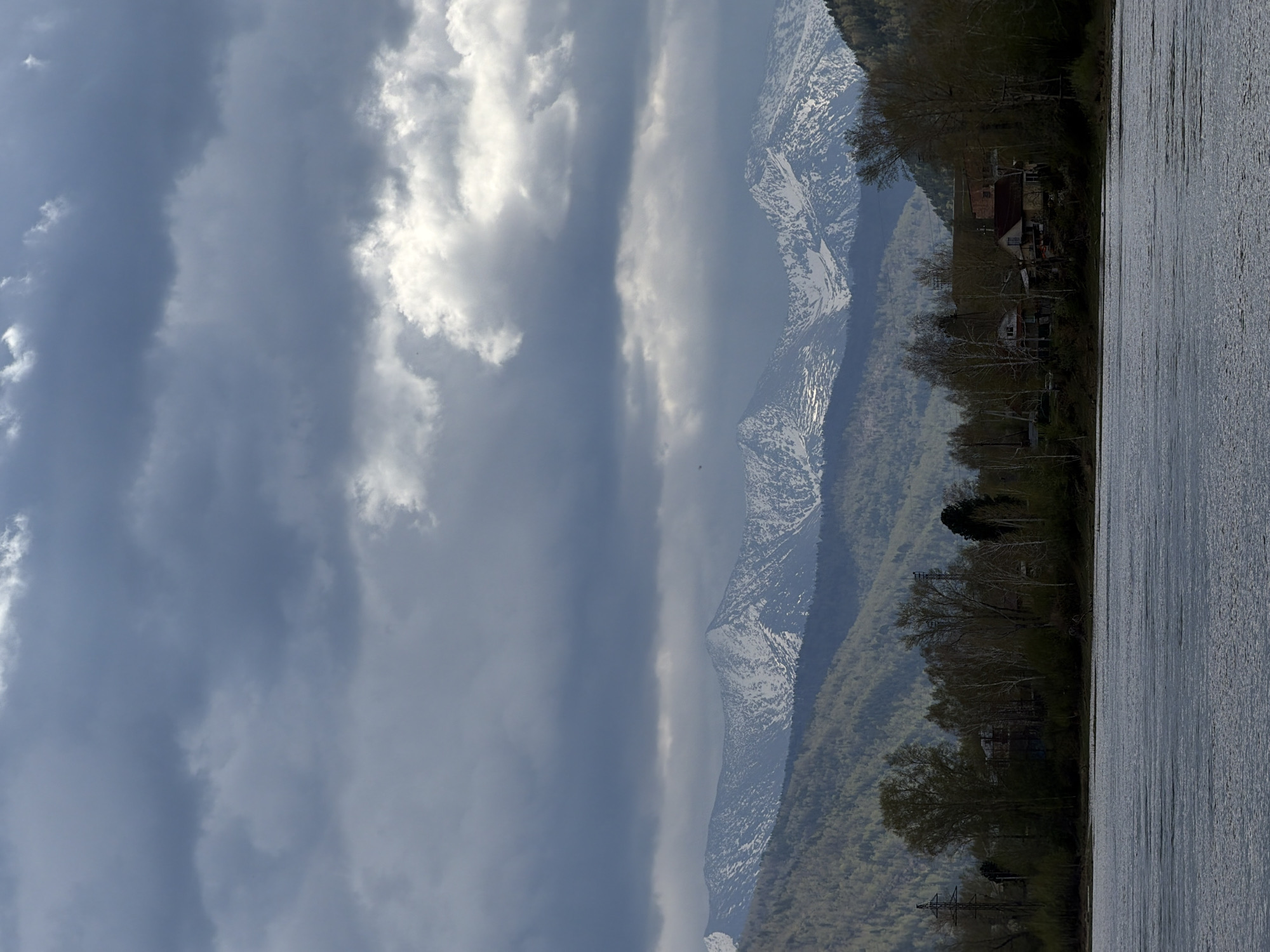 The image depicts a view of mountainous terrain near the city of Сизая, Russia. Snow-capped mountains are visible in the center of the frame, contrasted against a cloudy sky to the left. A body of water, possibly a lake or river, is on the right. The land near the water's edge has trees, houses, and utility poles.