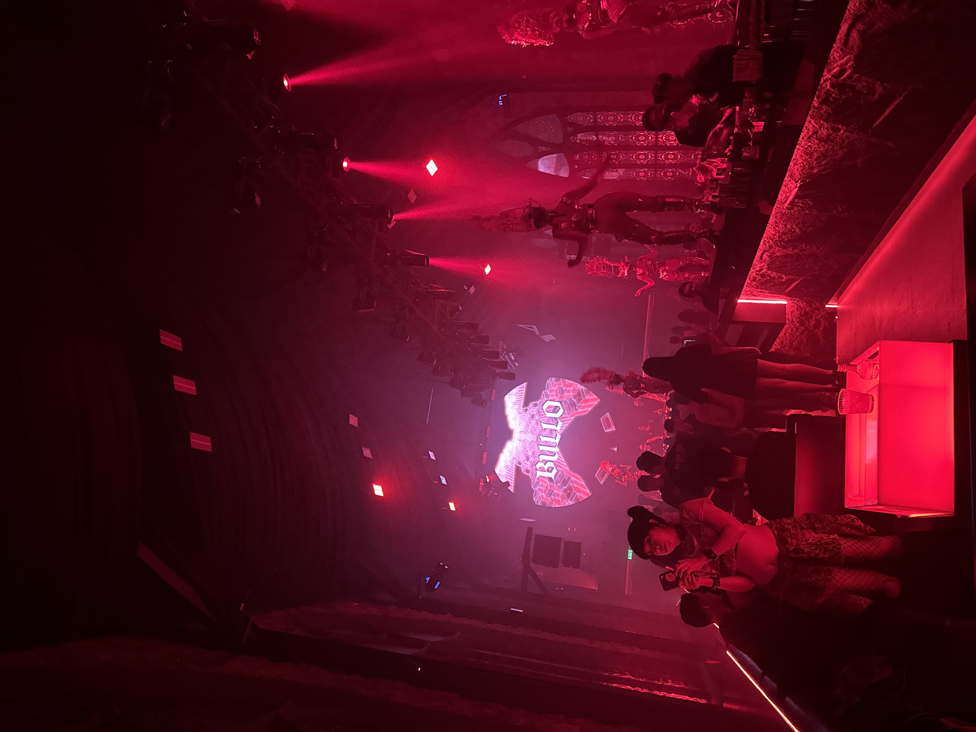 The image depicts the interior of a nightclub in North Kuta, Indonesia, bathed in vibrant red lighting. In the foreground, a woman with dark hair pulled into a bun is standing, holding a smartphone up to take a photo. She is wearing a crop top, fishnet stockings, and a patterned skirt. To her right, a glowing red table holds a drink. Behind her, other patrons are visible, some standing and some seated.
The midground of the image features a bar area with bartenders serving drinks. Above the bar, performers in elaborate costumes are dancing on elevated platforms, illuminated by bright red spotlights. A large screen in the background displays the word "BULLO" in a stylized font, framed by a graphic resembling a bowtie or wing-like structure. The overall ambiance is energetic and theatrical, consistent with a nightclub entertainment venue. The architectural elements suggest a dimly lit space with decorative patterns on the walls, resembling stained glass windows, further enhanced by the red lighting. An "EXIT" sign is visible on the left side of the frame.