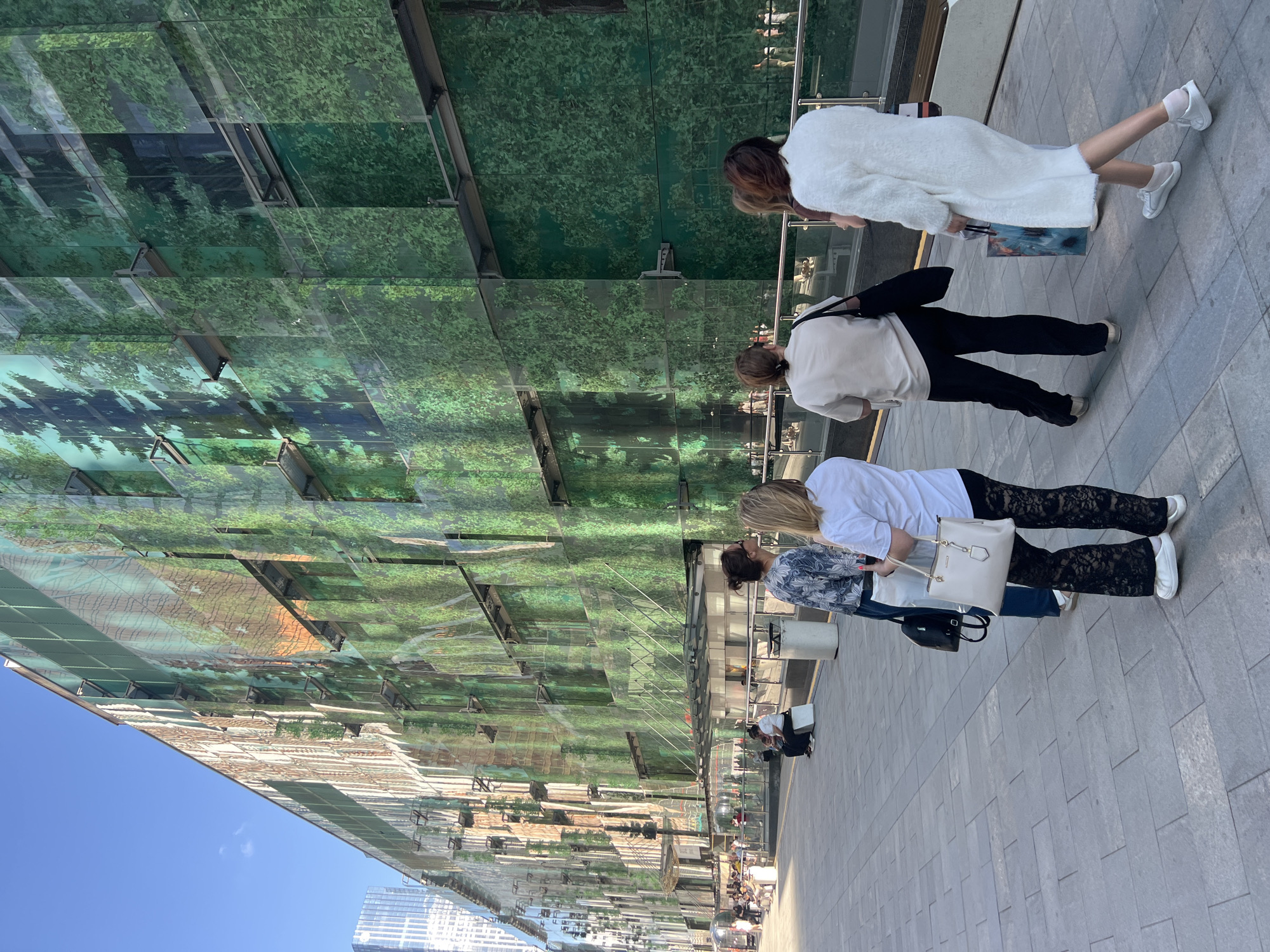The image depicts a street scene in Khimki, Russia, with a focus on a building facade adorned with a large-scale artistic print resembling a forest. The building's exterior is covered in green and brown panels, creating a camouflage-like effect with images of trees and foliage. A clear blue sky with a few wispy clouds is visible above.
In the foreground, four women are walking away from the camera on a paved walkway. The woman on the far left is partially visible and wearing a white top. The next woman is wearing a blue and white patterned shirt, dark pants, and carrying a white handbag. She is also wearing black lace pants. The third woman from the left is wearing a cream-colored t-shirt and black pants, carrying a black tote bag. The woman on the far right is wearing a long, fuzzy white coat and white sneakers, carrying a shopping bag.
Further in the background, several other people are visible sitting on benches or walking. The environment appears to be an urban setting with modern architecture. The overall impression is a sunny day in a contemporary city, with a notable artistic element on the building.