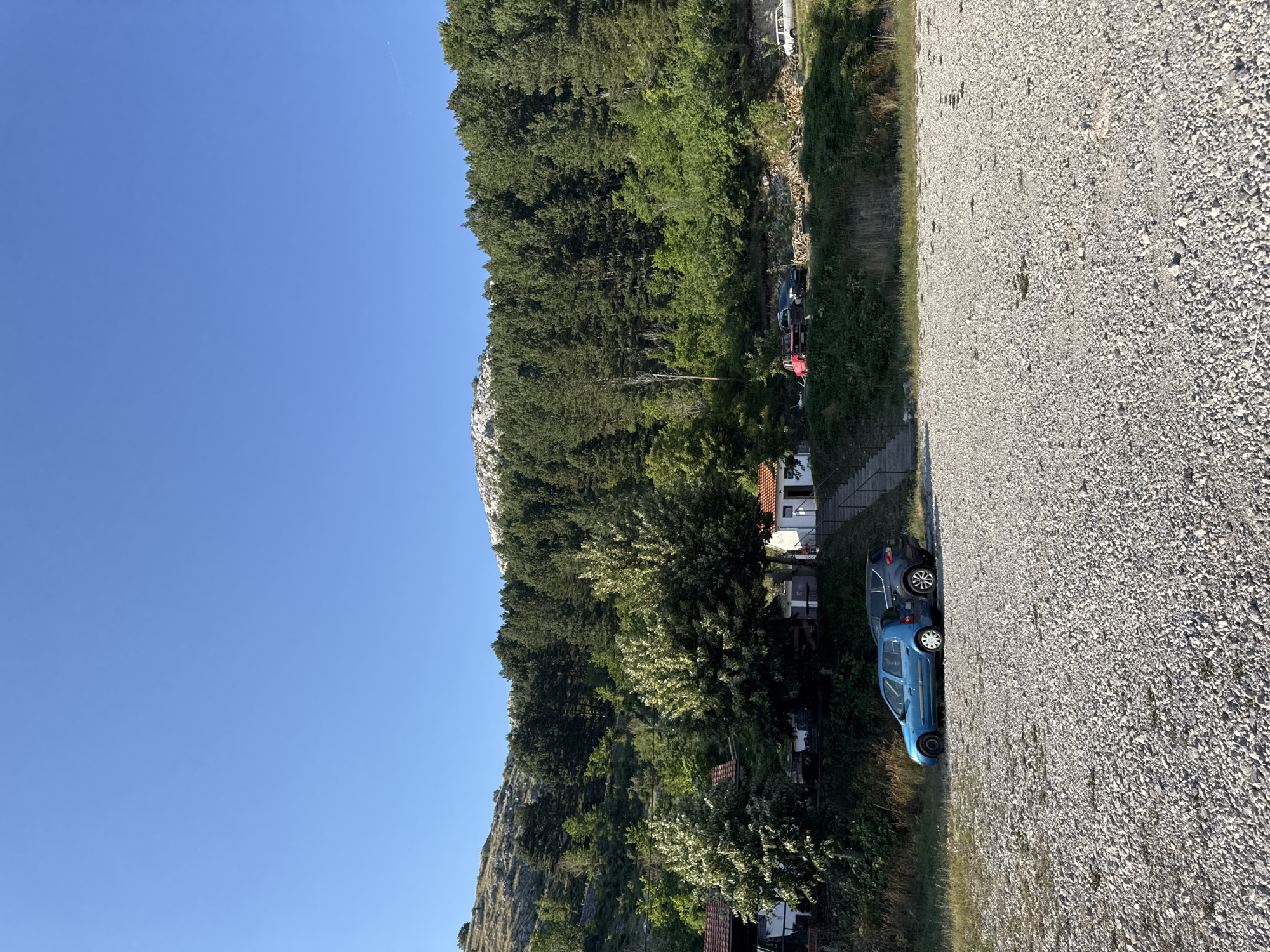 The image depicts a gravel parking area in front of a wooded hillside and a clear blue sky. In the foreground, two cars are parked on the gravel: a blue hatchback and a dark-colored SUV. Behind them, a small white building with a red roof is visible, with stairs leading up to its entrance. Further up the hill, a dense forest of pine trees covers the landscape, leading to a rocky, mountainous peak in the distance. Several other vehicles, including a red pickup truck and a white van, are parked along a road higher up on the hillside. The scene appears to be a remote location, possibly in a rural area like Bukovica, Montenegro, given the natural surroundings and the presence of a small dwelling and parking area.