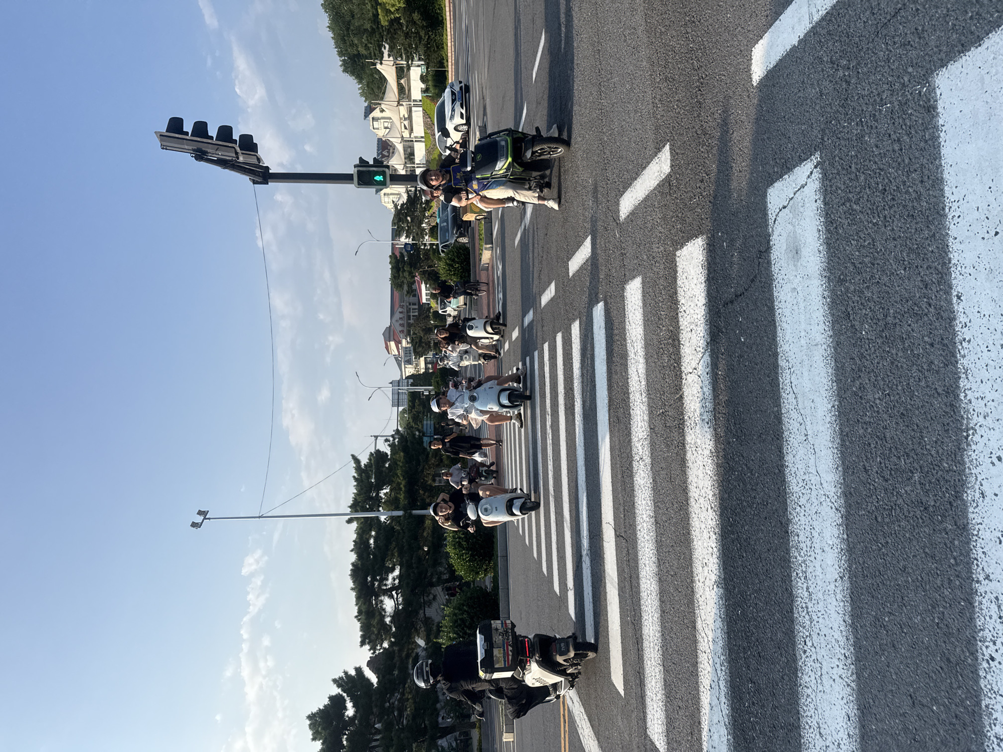 The image depicts a street scene in Qinhuangdao, China, likely near the coast given the presence of lush greenery and a clear sky. Several people are riding electric scooters and motorcycles on a paved road with a pedestrian crossing. The traffic light is green, indicating that traffic is allowed to proceed. In the foreground, a person on a black scooter is approaching the pedestrian crossing, followed by others on similar vehicles. In the background, more scooters and a few cars are visible. The overall atmosphere suggests a typical urban environment during daylight hours, with clear weather conditions. The architecture in the background features modern buildings with white awnings, hinting at a commercial or residential area.