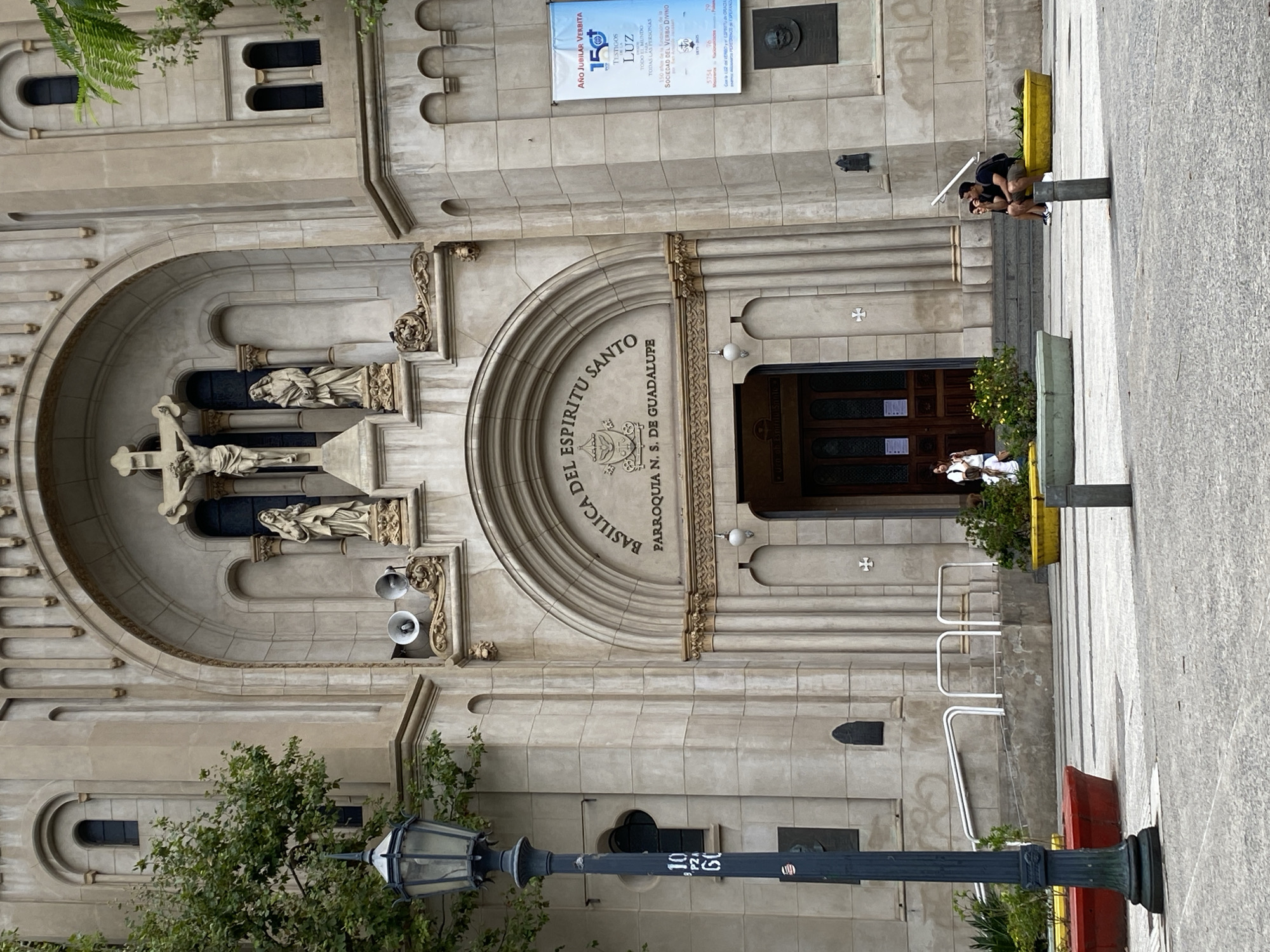 An eye-level exterior view captures the facade of the Basilica Nuestra Señora de Guadalupe, located at Paraguay 3901, Buenos Aires, Argentina, during daytime. The basilica's light-colored stone or stucco facade features classical and Romanesque architectural elements, including arched windows and recessed panels. The central main entrance consists of double wooden doors beneath a prominent arch, inscribed with "BASILICA DEL ESPIRITU SANTO PARROQUIA N. S. DE GUADALUPE". Above this, within a large arched niche, a crucifixion sculpture depicts Christ on the cross, flanked by two standing statues. Two white loudspeakers are mounted below this niche. To the right of the entrance, a vertical white banner displays "AÑO JUBILAR VERBITA 150" and other text, with a circular relief portrait affixed below it. Graffiti is present on the lower left facade.
In the foreground, a dark blue/black ornate lamppost with an oval lantern stands on the left, marked "10 PZ 60". Green tree foliage is visible behind it and to the far right. The ground is a patterned gray paved area. Red, yellow, and green rectangular and circular planters containing green plants are arranged on the pavement. White metal railings provide access to the basilica's entrance via concrete steps. A dark gray cylindrical bollard is in the mid-ground.
Four individuals are present. Two individuals are seated on a yellow planter to the right: a male in a dark t-shirt and shorts holds a white open umbrella overhead, while a female in a dark top and shorts sits beside him. Near the main entrance, a female in a white top and dark pants stands in the doorway, and another female in a white top and light pants sits on the steps to the left of the entrance.