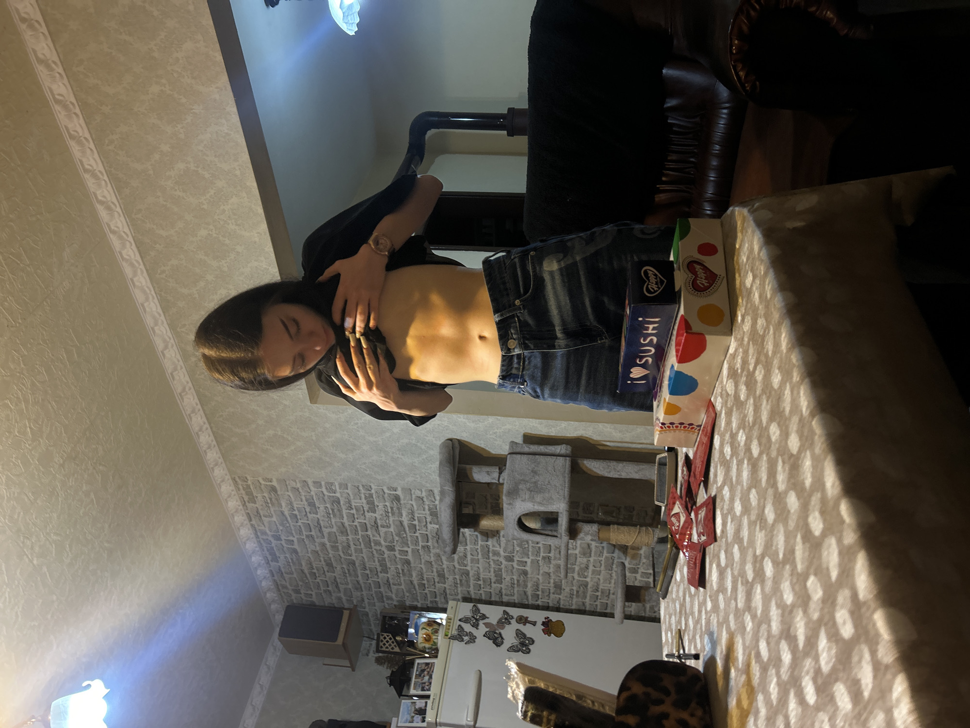 The image shows a woman indoors, wearing dark jeans and a black top that is pulled up to expose her abdomen. She is standing in a room with wallpaper and a brick-patterned wall. A cat tree and a white refrigerator with butterfly decorations are visible in the background. There is a table with a patterned tablecloth in the foreground, with boxes of sushi and other items on it. The location is likely a home in Burgas, Bulgaria, but there are no specific landmarks that confirm this.