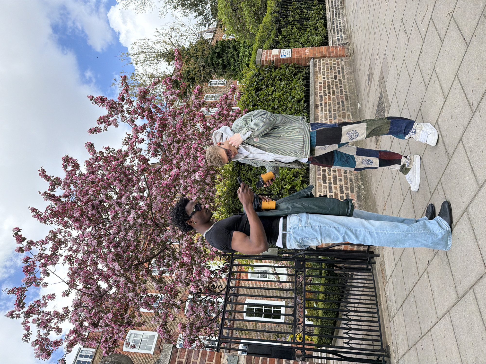 Two young men are standing on a paved sidewalk in a residential area, likely in London, United Kingdom. The man on the left is Black, wearing a black t-shirt, light blue jeans with a white belt, black shoes, and sunglasses. He is holding a smartphone in one hand and what appears to be a yellow cup in the other. A long, dark green object is strapped across his torso. He is standing in front of a black iron gate.
The man on the right is white, wearing a striped green and white jacket over a grey hoodie, patchwork pants, and white sneakers. He has his hand near his face and is looking at the other man.
Behind them are brick buildings with white-framed windows, lush green foliage, and a tree with pink blossoms. The sky is visible with blue patches and white clouds.