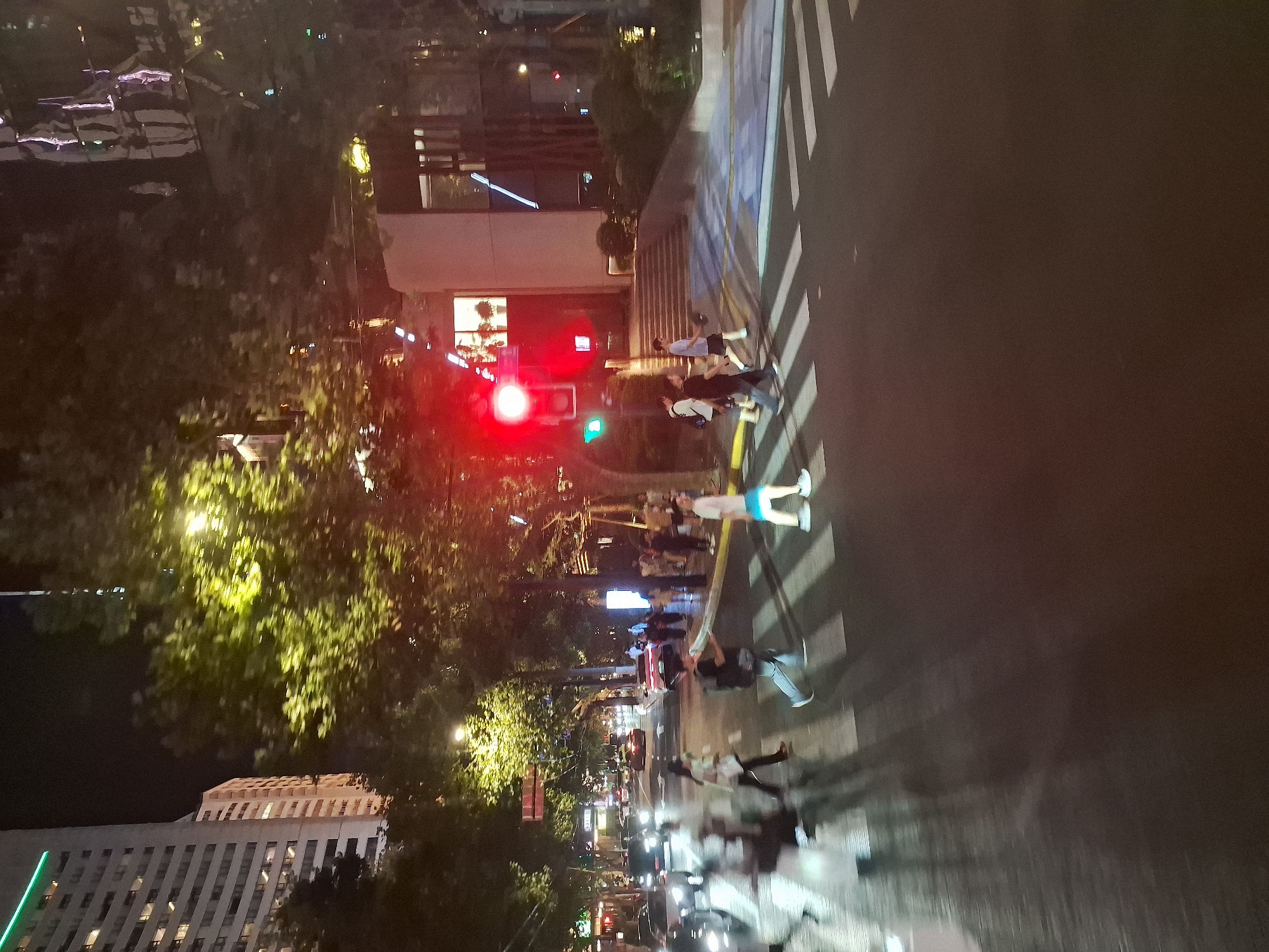 This image captures a night scene in Shanghai, China, likely along a busy street with pedestrian activity. The foreground shows a crosswalk with several people crossing. Motion blur is evident, indicating movement and possibly a low light photography setting or a moving camera. To the left, a tall, modern building with illuminated windows is visible, suggesting an urban environment. Trees line the street, their leaves illuminated by streetlights, creating a dappled lighting effect. Traffic lights are present, with a red light clearly visible. In the background, other vehicles and buildings contribute to the city atmosphere. The overall impression is one of a bustling city at night, with people navigating the urban landscape.