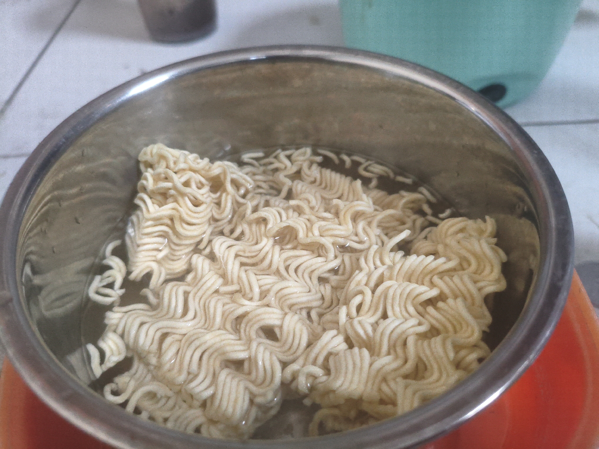 A close-up, overhead view captures a stainless steel pot filled with instant noodles submerged in clear water. The noodles are dry and corrugated, stacked in a compact block. The pot rests on an orange surface, possibly a placemat or a pot holder, with a blurred background showing a white tiled surface and a partial view of a light blue appliance. The lighting is dim and diffused, contributing to a grainy texture throughout the image. No people or specific locations within Wujil, Indonesia are identifiable.